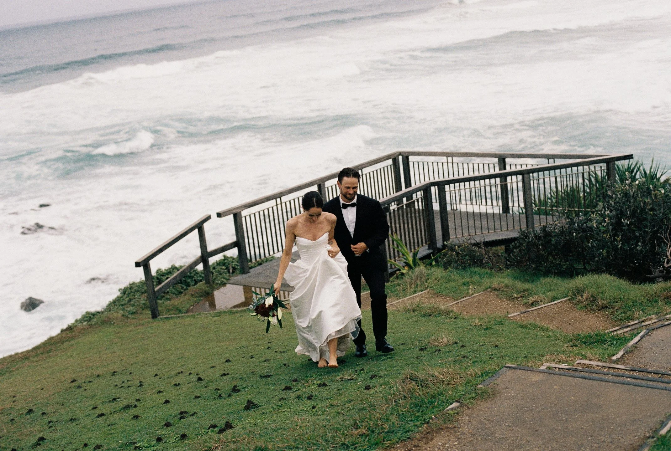 A bride and groom walking barefoot on grass near the beach, with the ocean waves in the background and a wooden railing along the path.