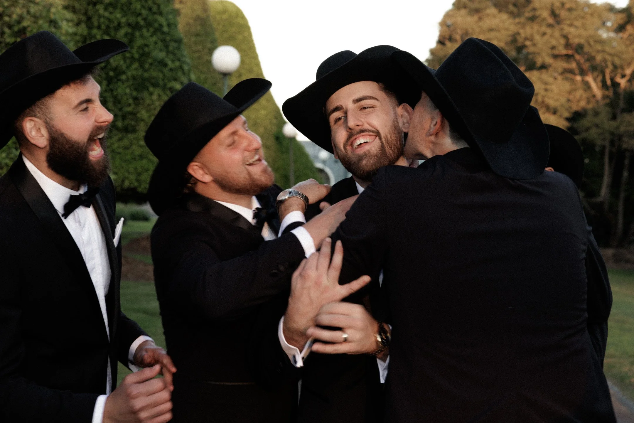 Men wearing black tuxedos and wide-brimmed black hats celebrating and hugging outdoors during sunset, with greenery and lampposts in the background.
