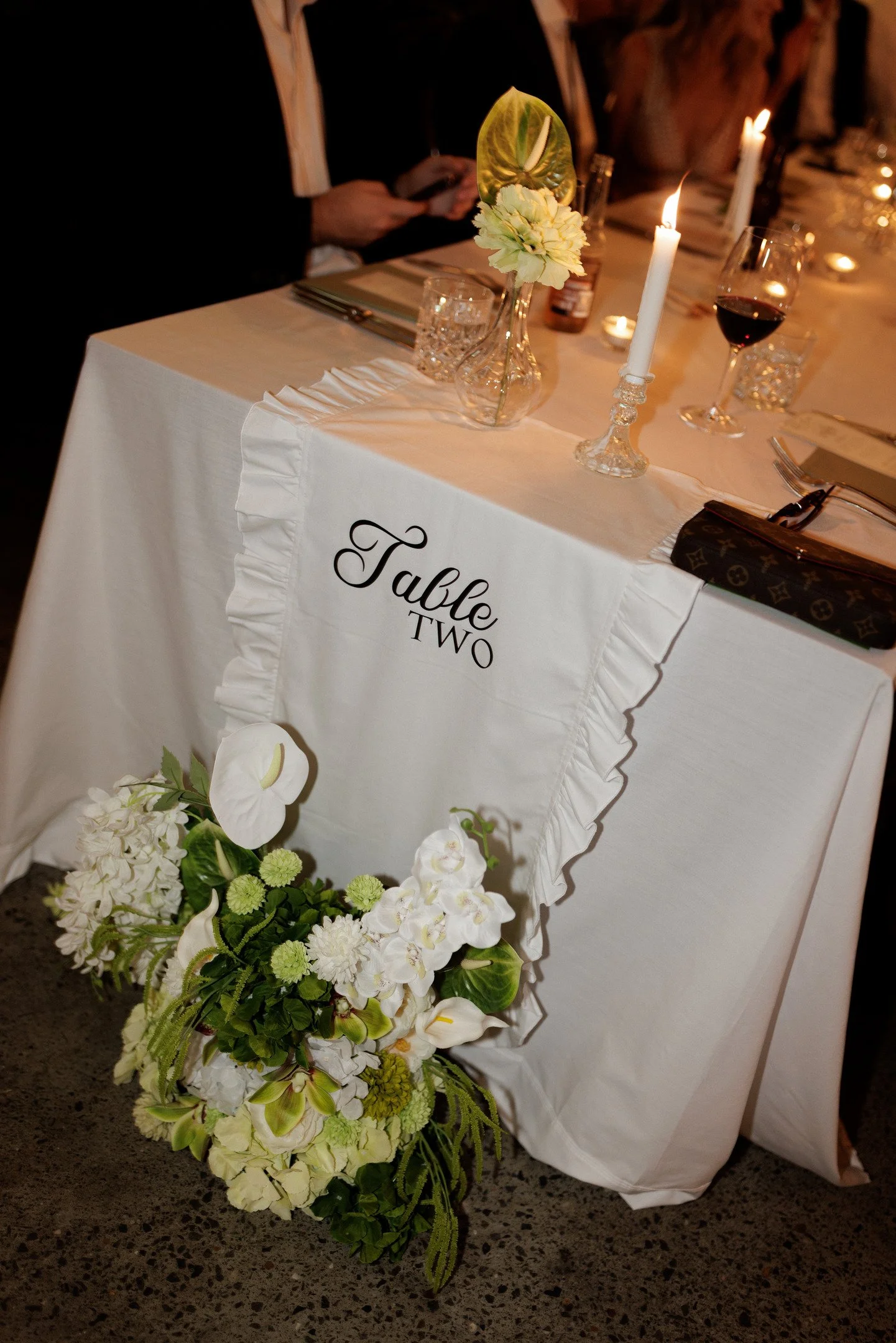 A decorated table with white tablecloth, floral arrangement, white candles, and wine glasses, labeled 'Table Two,' with a person using a smartphone in the background.