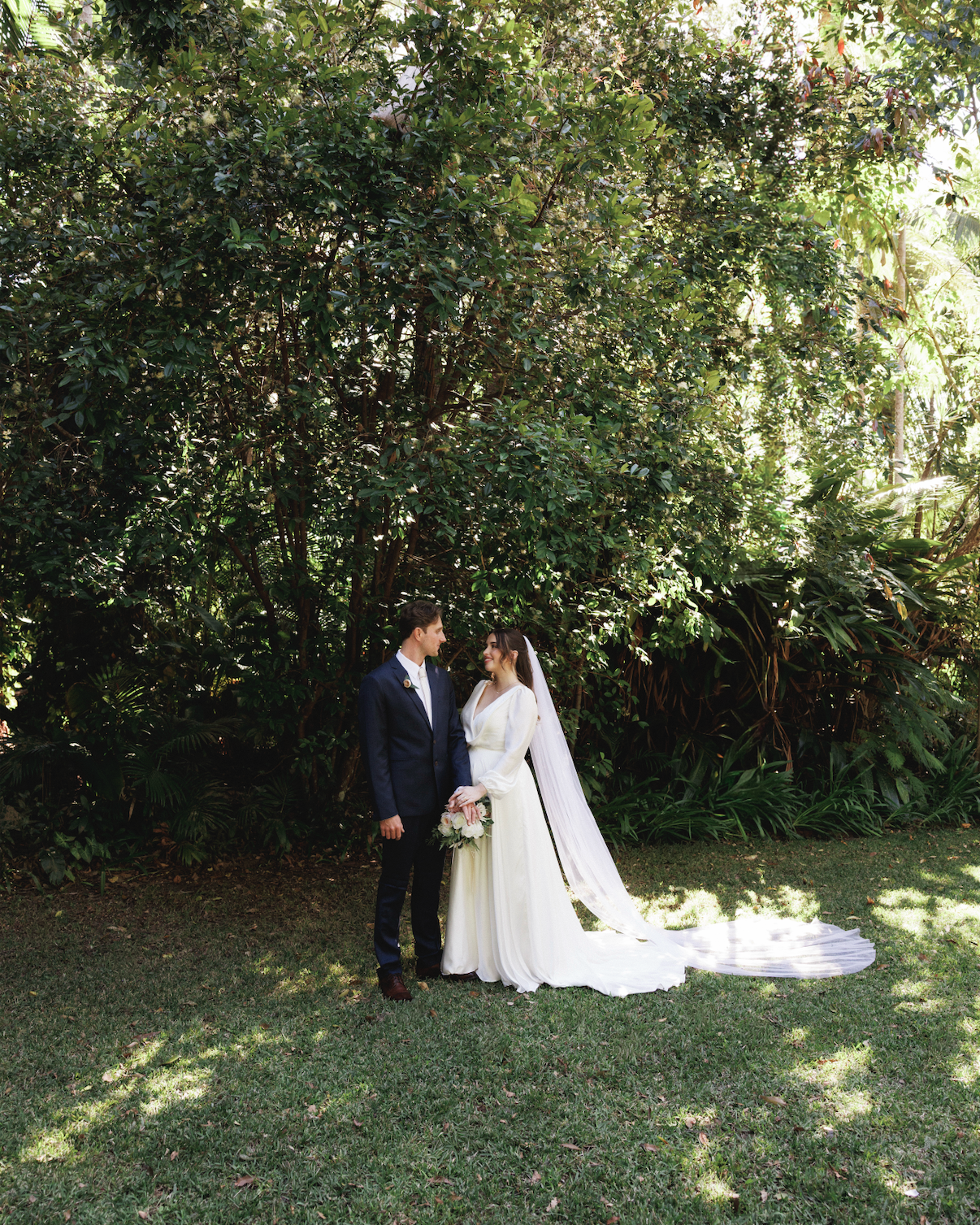 A bride and groom standing outdoors in front of dense green foliage, holding hands and looking at each other, with the bride holding a bouquet of flowers.