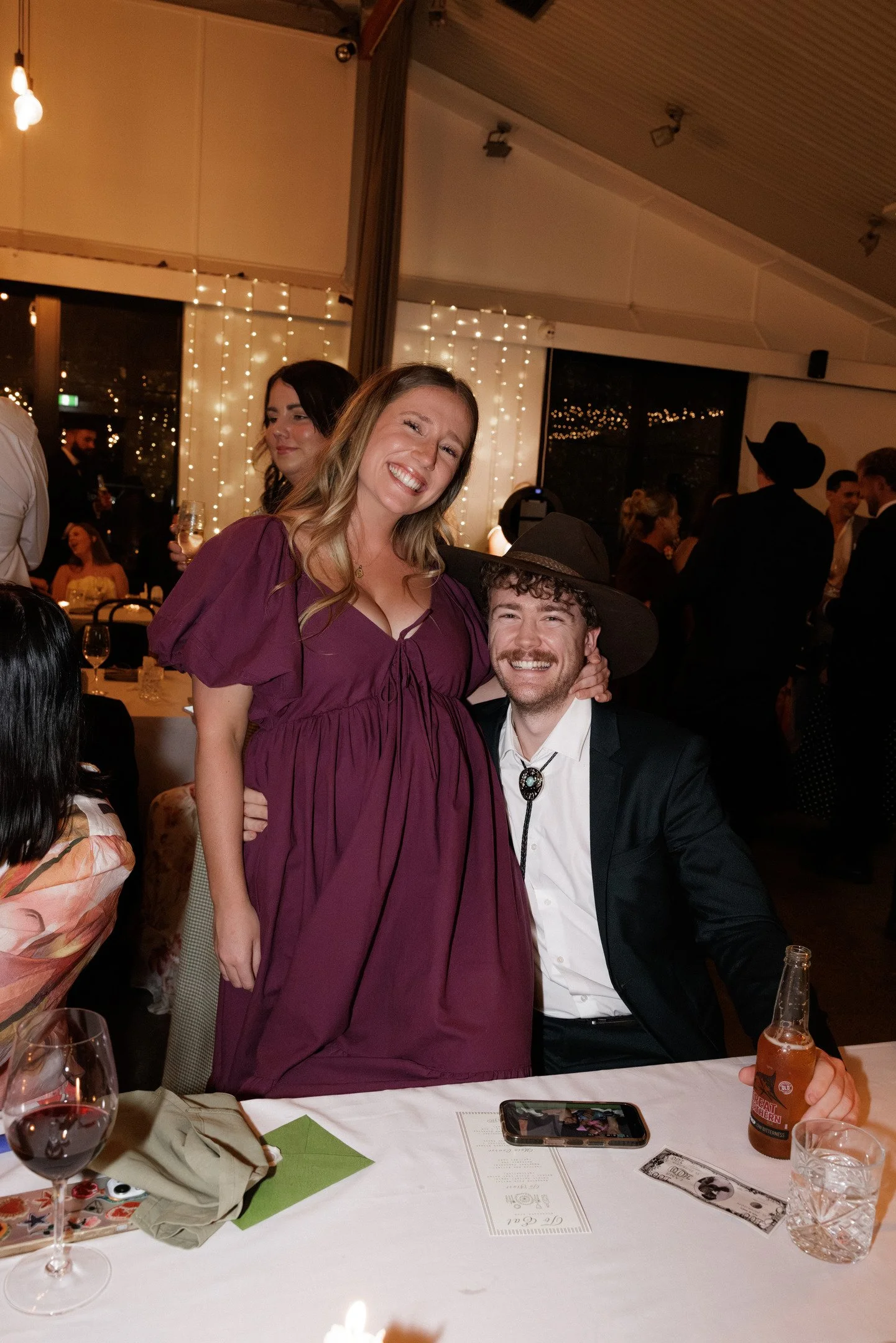A woman in a purple dress smiling and standing next to a man in a cowboy hat sitting at a dinner table during a celebration or party in a decorated event space at night, with guests in the background.