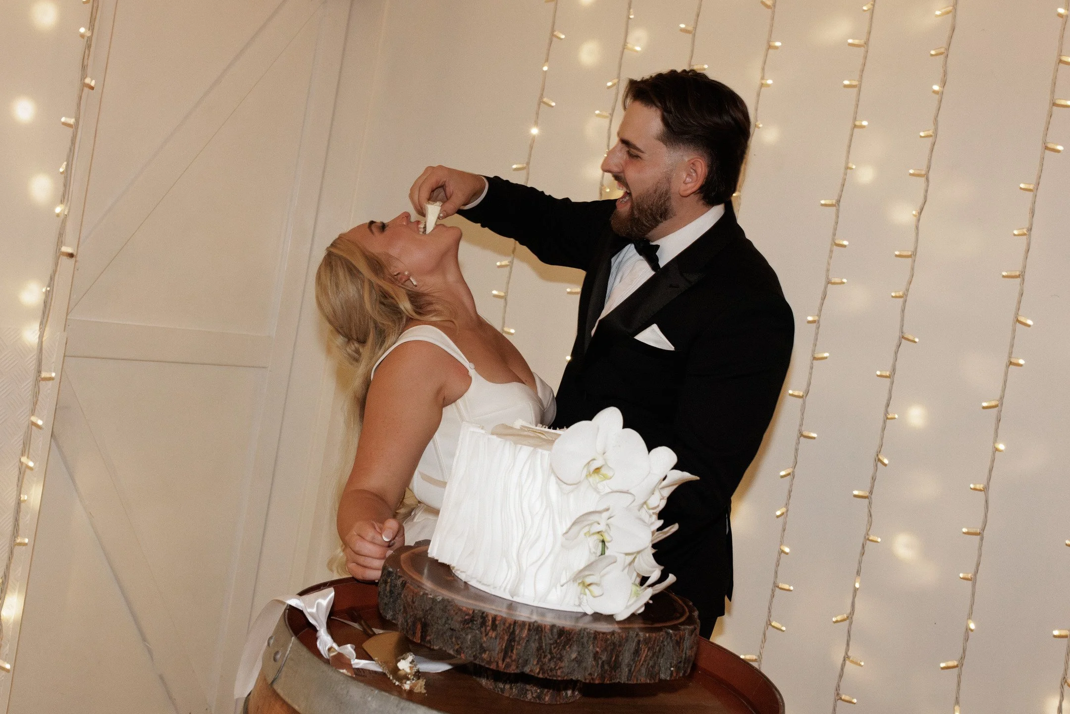 A man and woman in formal attire cutting a wedding cake together, with the woman feeding cake to the man, at a celebration event.