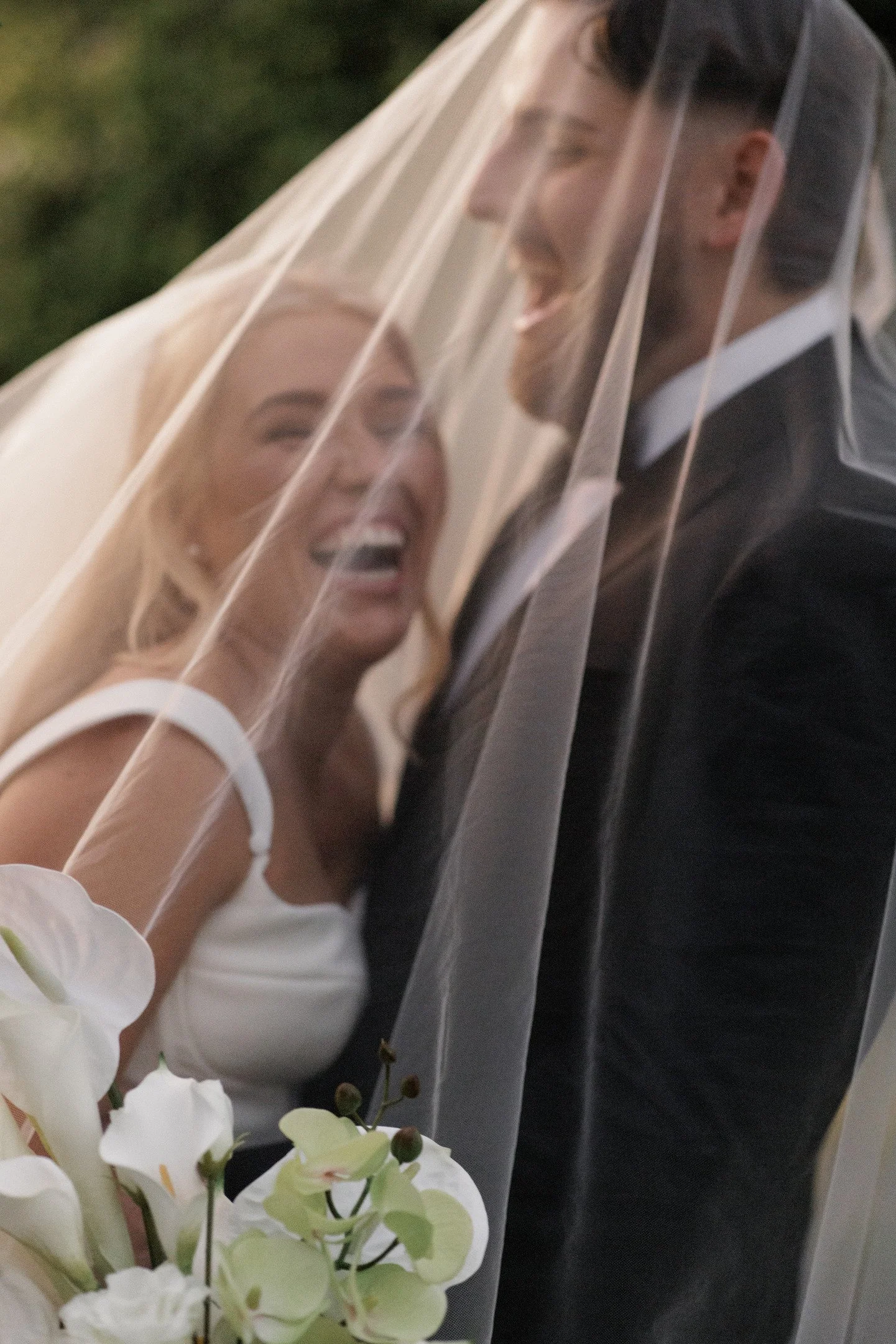 A smiling bride and groom behind a wedding veil, with white orchids in the foreground.