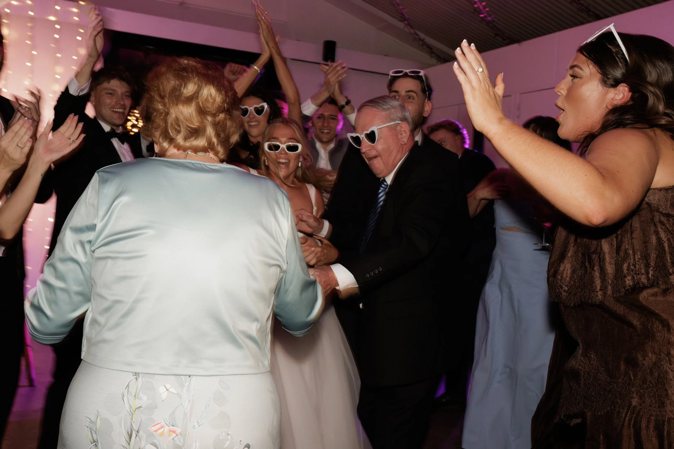 Group of people at a celebration or party, wearing formal attire and sunglasses, dancing and smiling.