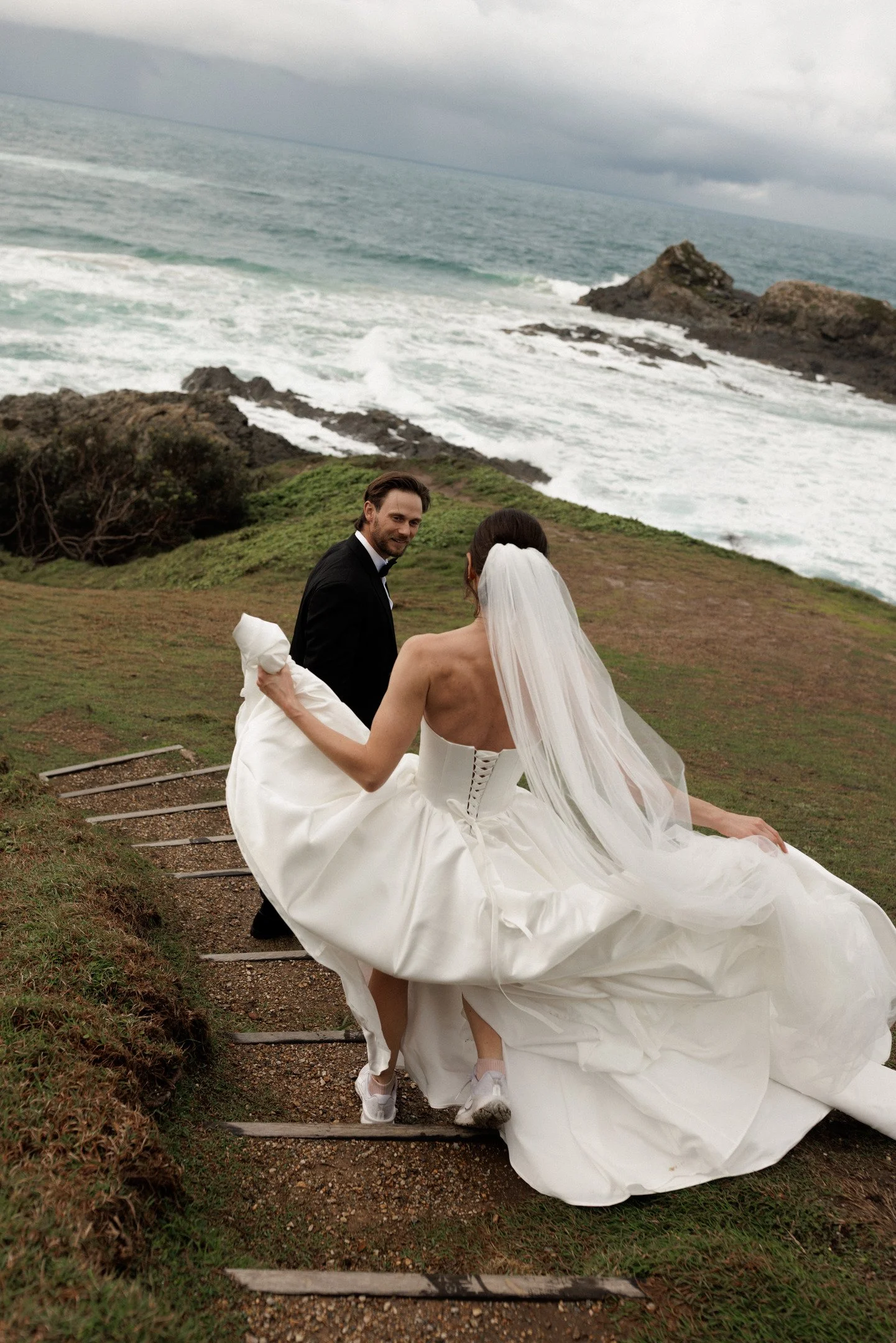 A couple dressed in wedding attire near the coast, with rocky cliffs and the ocean in the background under a cloudy sky.