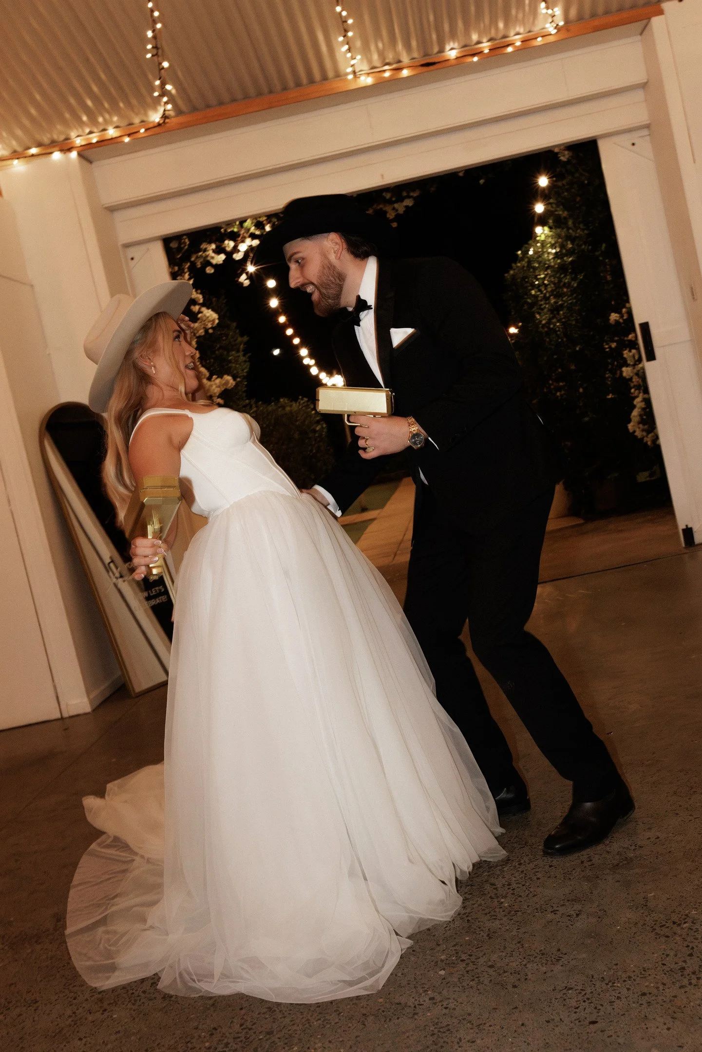 A woman in a white wedding dress and hat smiling at a man in a black tuxedo and cowboy hat during a wedding celebration, with string lights overhead and a dark outdoor background.