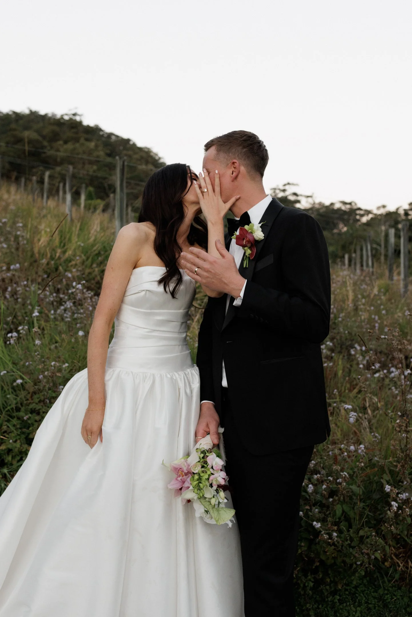 A bride and groom kiss outdoors, in wedding attire, with greenery and flowers in the background.