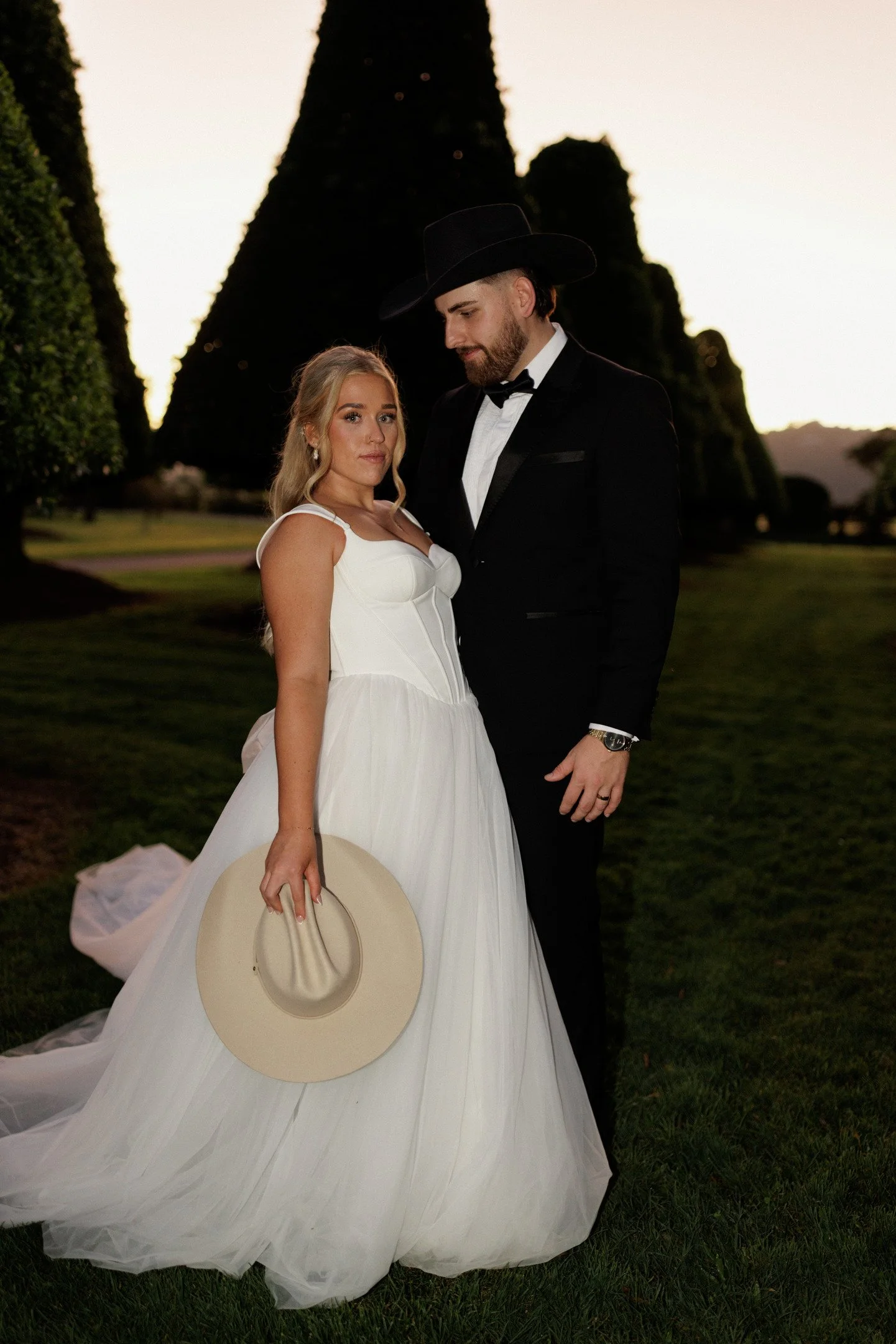 A bride and groom standing outdoors during sunset. The bride is wearing a white wedding dress and holding a wide-brimmed hat, while the groom is dressed in a black tuxedo with a bow tie and a black hat.