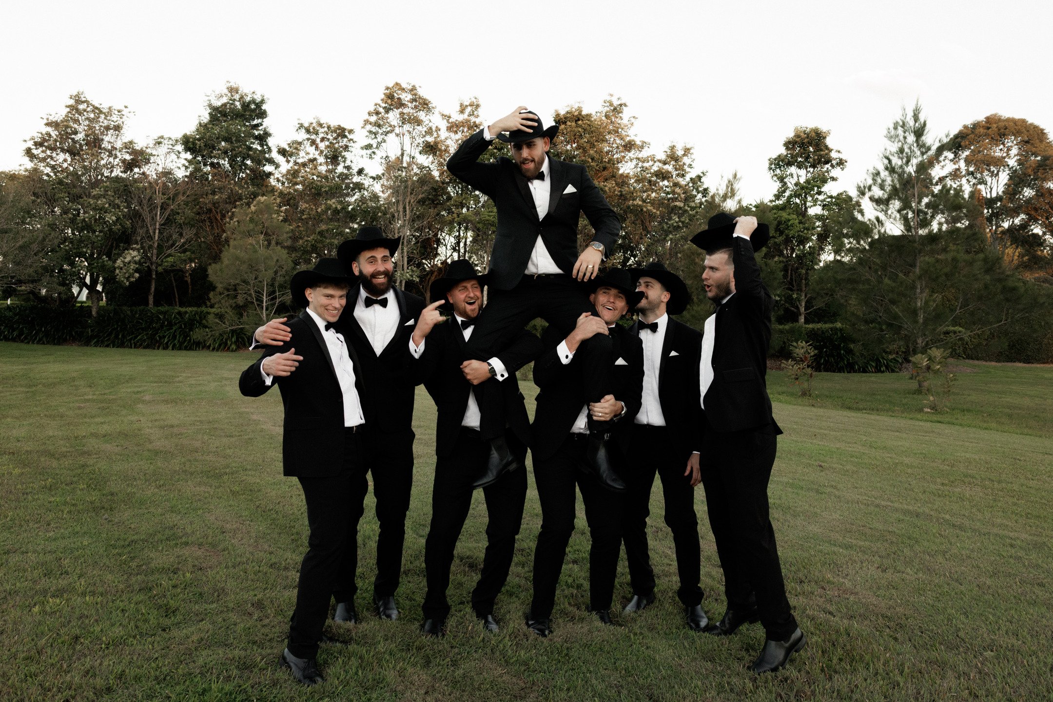 A group of men dressed in black tuxedos and cowboy hats, forming a circle outdoors on a grassy field with trees in the background. One man is lifted in the center, holding his hat, with others supporting and smiling.