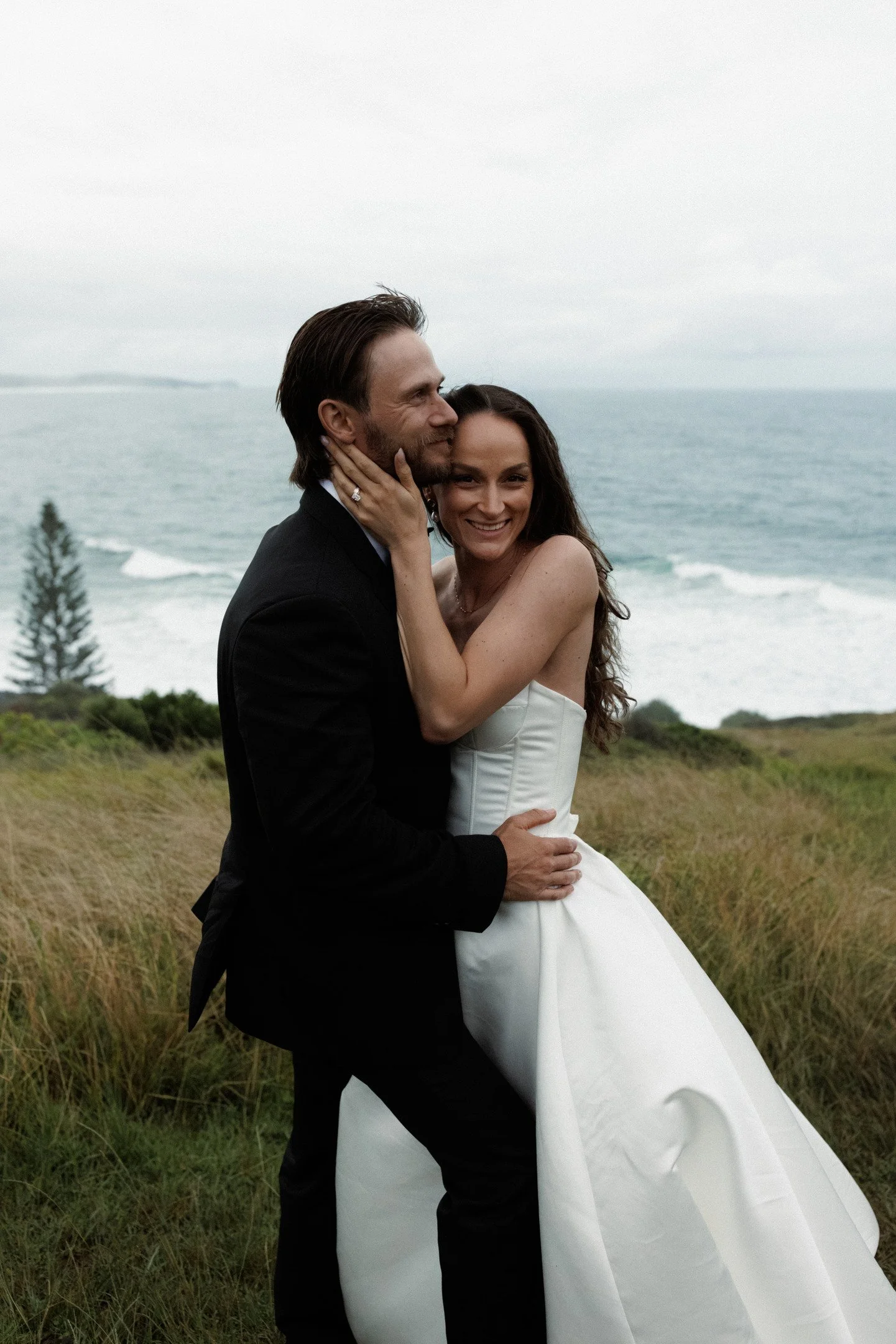 A newlywed couple embracing outdoors with the ocean in the background, woman in a white wedding dress and man in a black suit.
