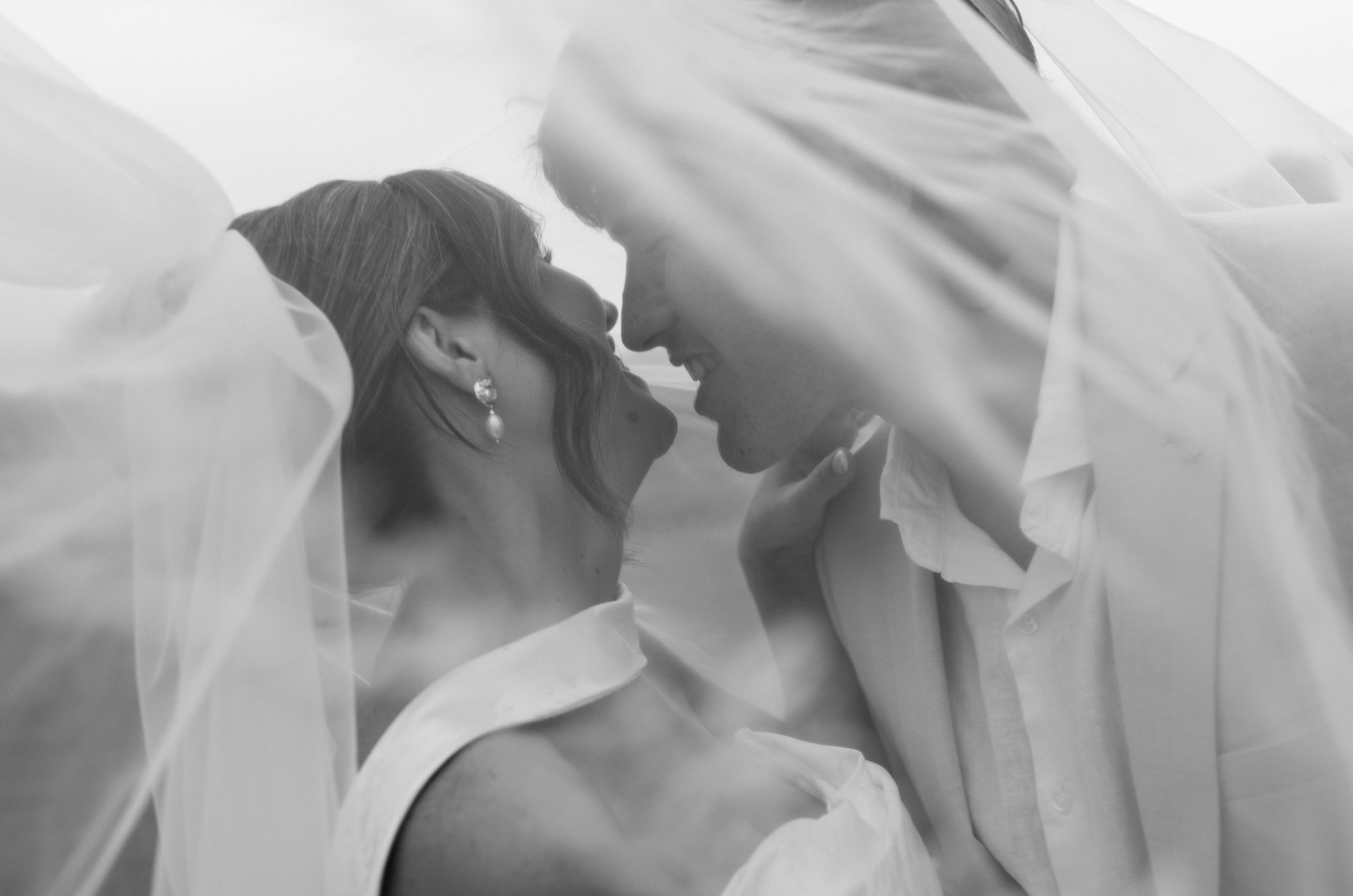 A couple on their wedding day, close together under a veil, smiling and touching foreheads, in black and white.