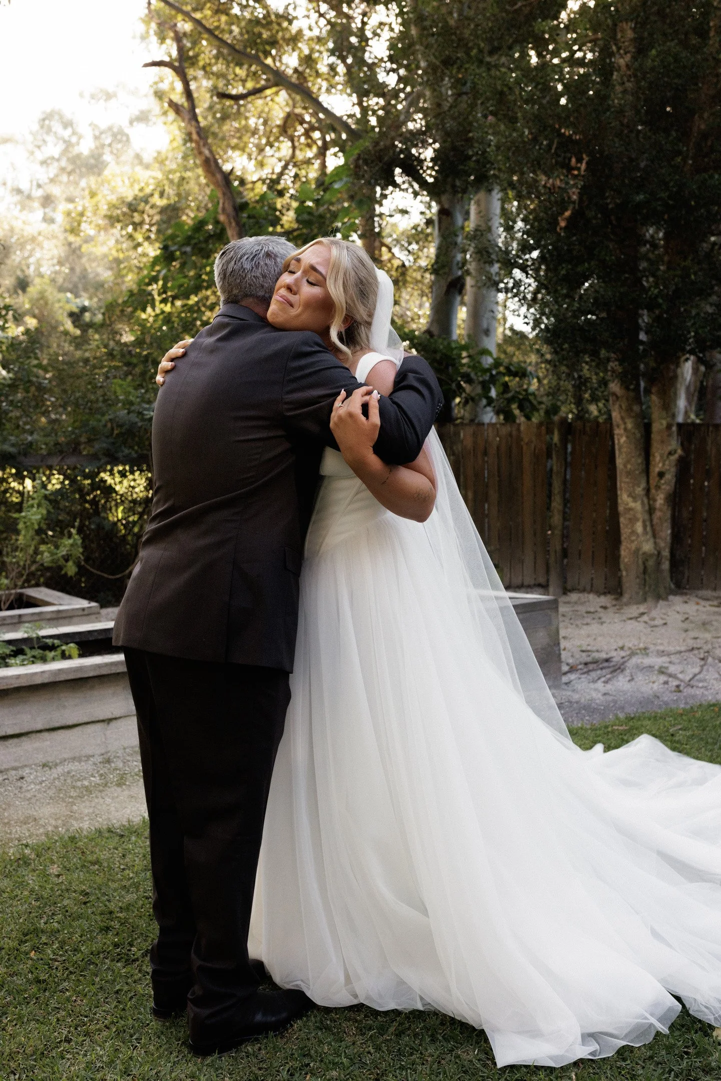 A woman in a white wedding dress is hugging a man in a black suit outdoors, with trees and a wooden fence in the background.