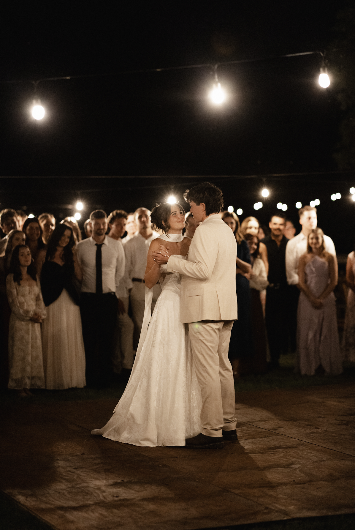 A bride and groom dancing at their wedding reception outdoors at night, surrounded by guests and illuminated by string lights.