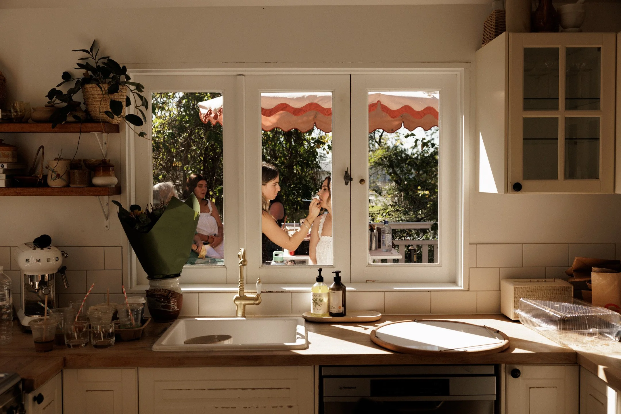View through a kitchen window showing three women outside, one applying makeup or lipstick to another, with trees and sunlight in the background.