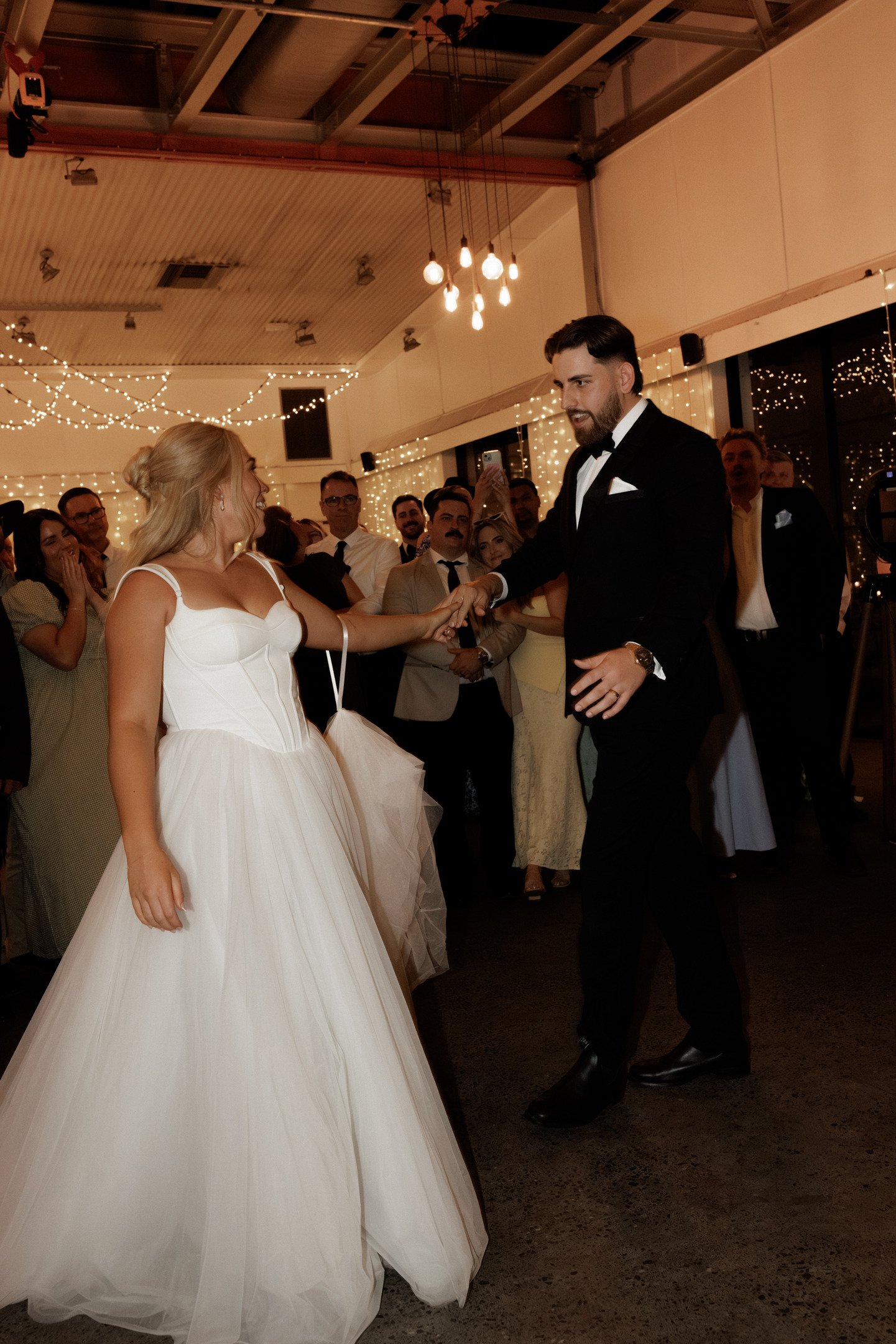 A bride and groom dancing at their wedding reception, surrounded by guests in a decorated indoor venue with string lights.