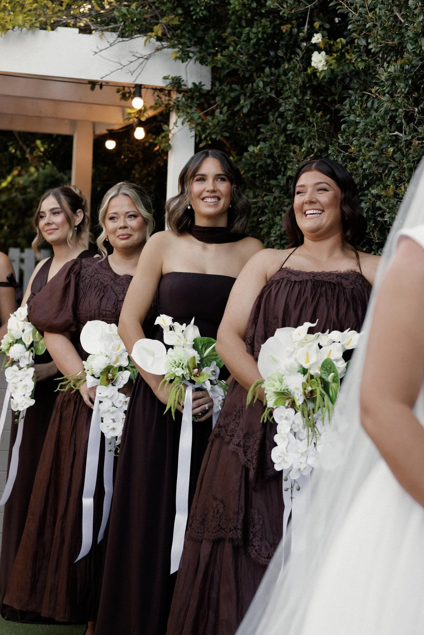 Four women in matching brown dresses holding white flower bouquets, standing outdoors during a wedding ceremony, with greenery and hanging lights in the background.