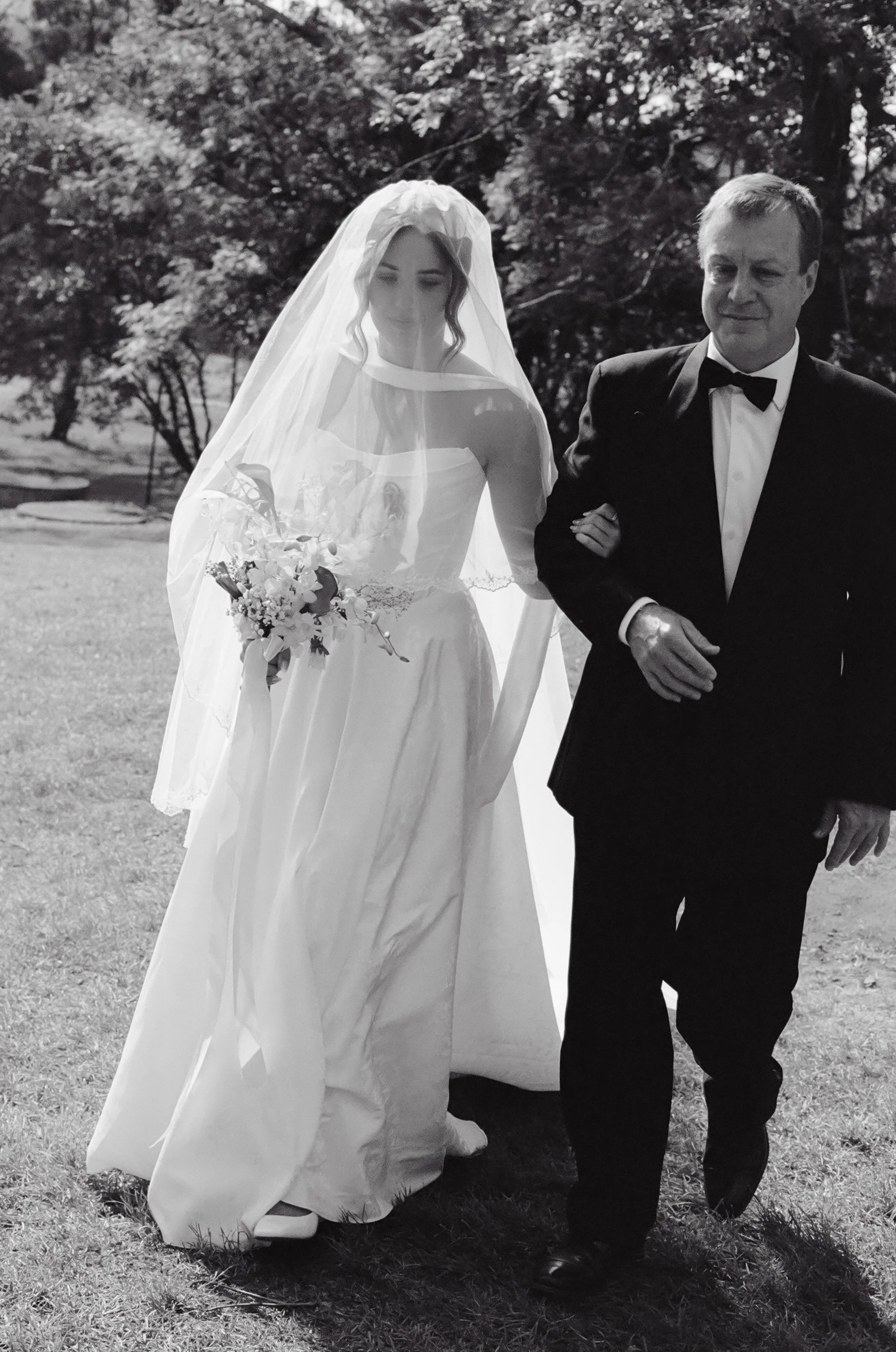 A bride in a white wedding dress with a veil and bouquet walking outdoors with an older man in a tuxedo.