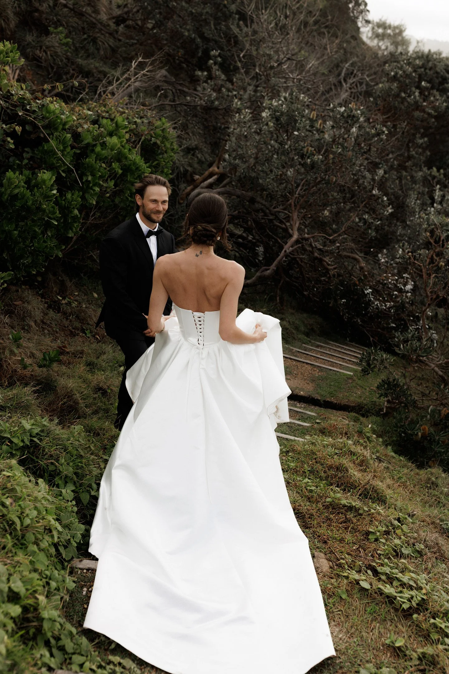 A man in a black tuxedo and bow tie holding hands with a woman in a white wedding gown outdoors in a natural setting with trees and greenery.