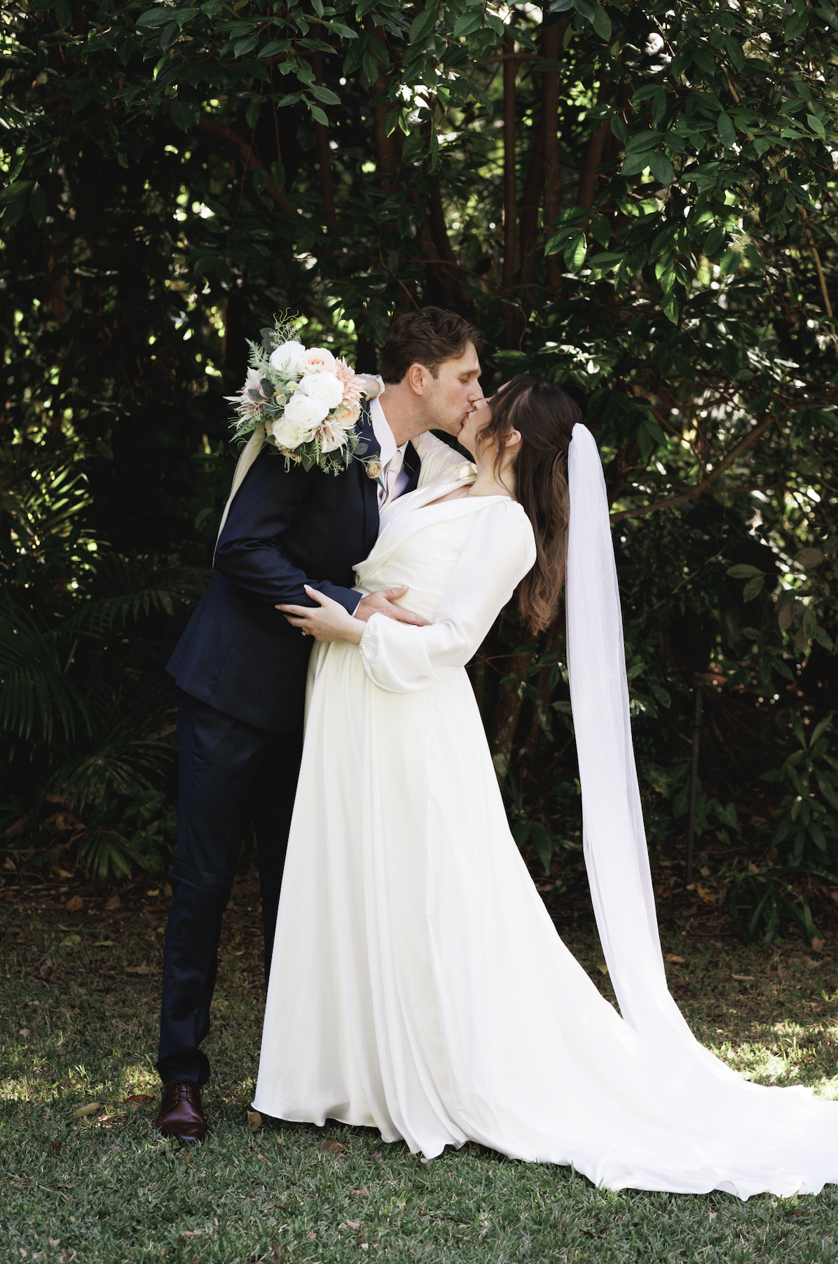 A newlywed couple sharing a kiss outdoors among green foliage. The groom holds a bouquet of white and pink flowers over his shoulder, and the bride is wearing a white dress with a long veil.