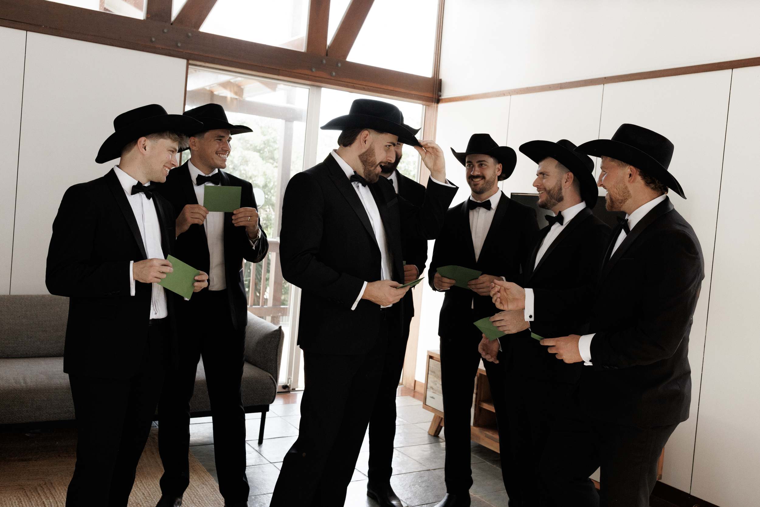 A group of seven men dressed in black tuxedos and black cowboy hats, standing indoors near a window, appearing to be engaged in a conversation or preparing for an event.