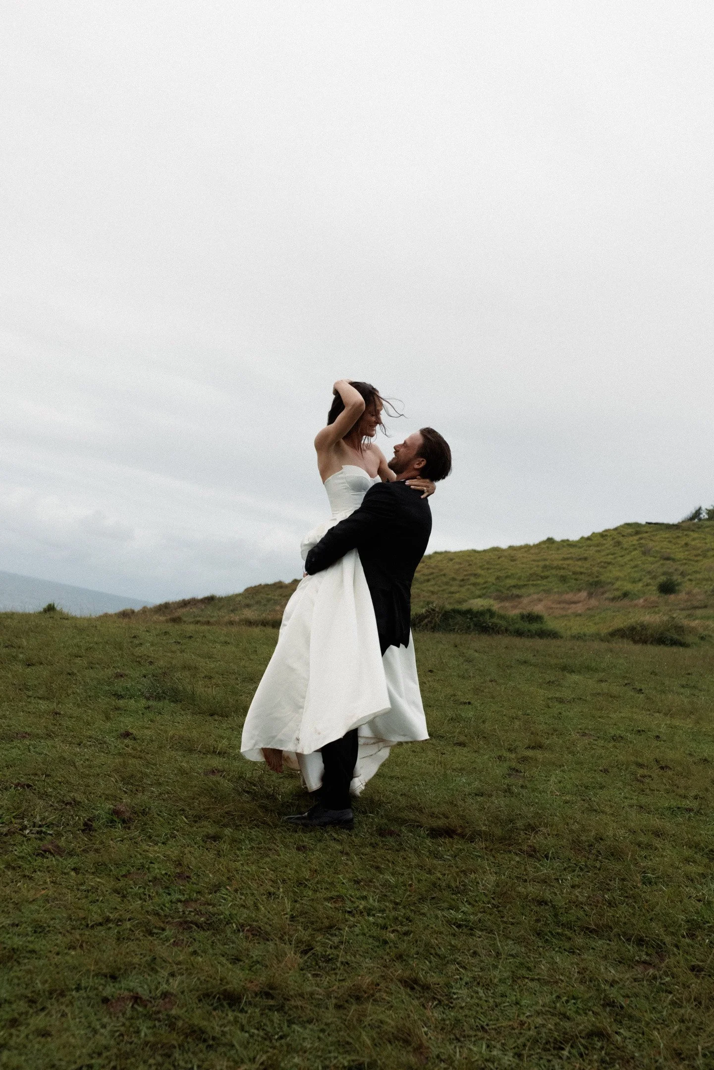 A man in a black suit and a woman in a white wedding dress are celebrating outdoors on a grassy hill with cloudy skies in the background, with the man lifting the woman in an embrace.