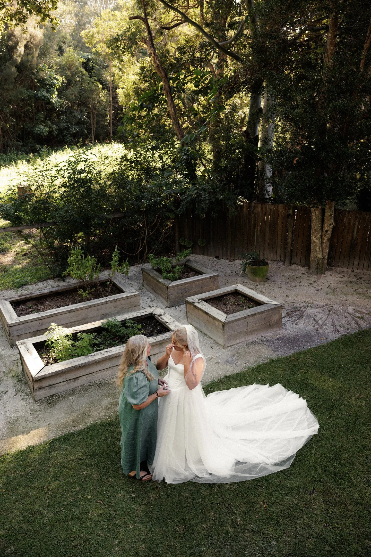 A bride and a woman in an outdoor garden setting, with the bride wearing a white gown and the woman in casual clothing, standing close together and holding hands, with garden beds and trees in the background.