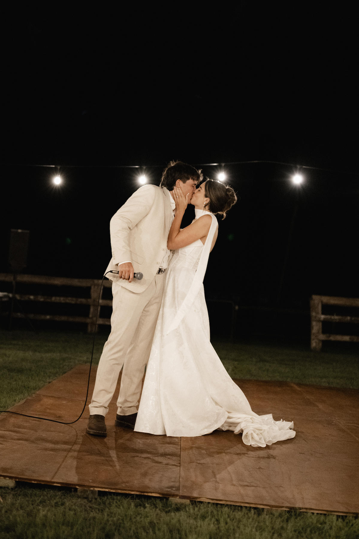 A couple in wedding attire sharing a kiss during their outdoor wedding reception at night, standing on a wooden platform with string lights overhead.