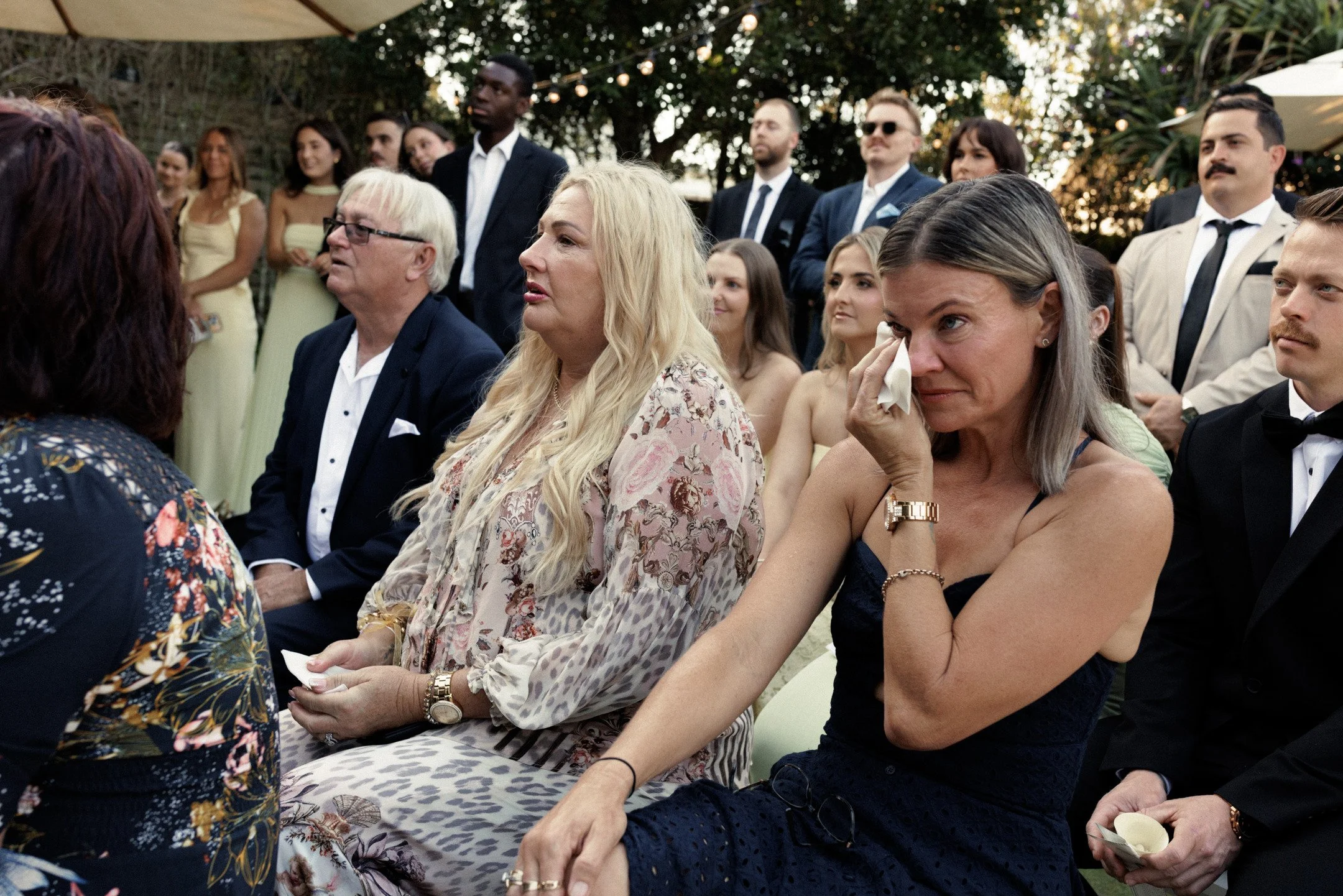 A group of people dressed in formal attire attending a gathering outdoors during the evening.