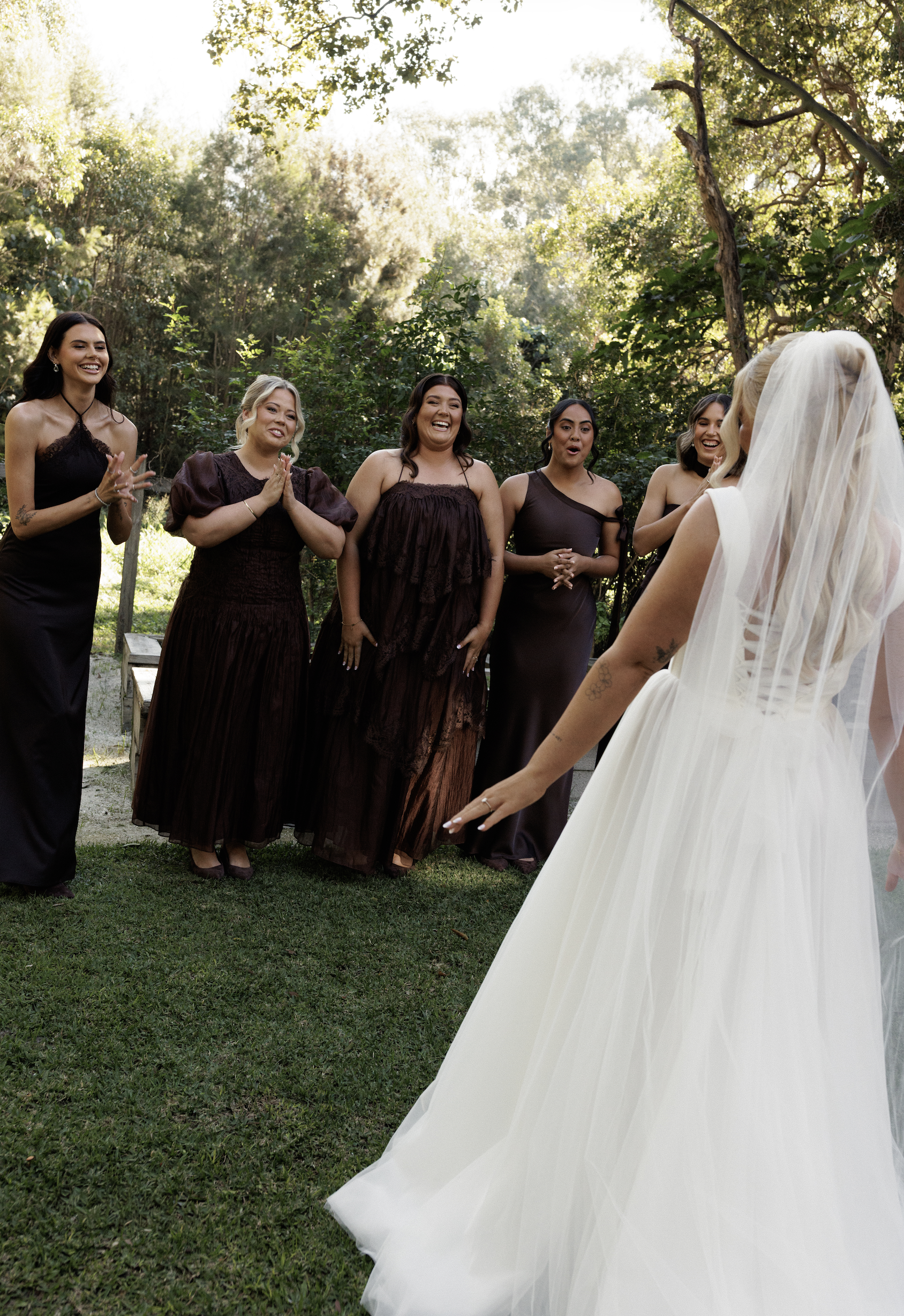A bride in a white wedding dress with a veil walks outdoors on grass, while five women in black and brown dresses watch and smile, with trees and sunlight in the background.
