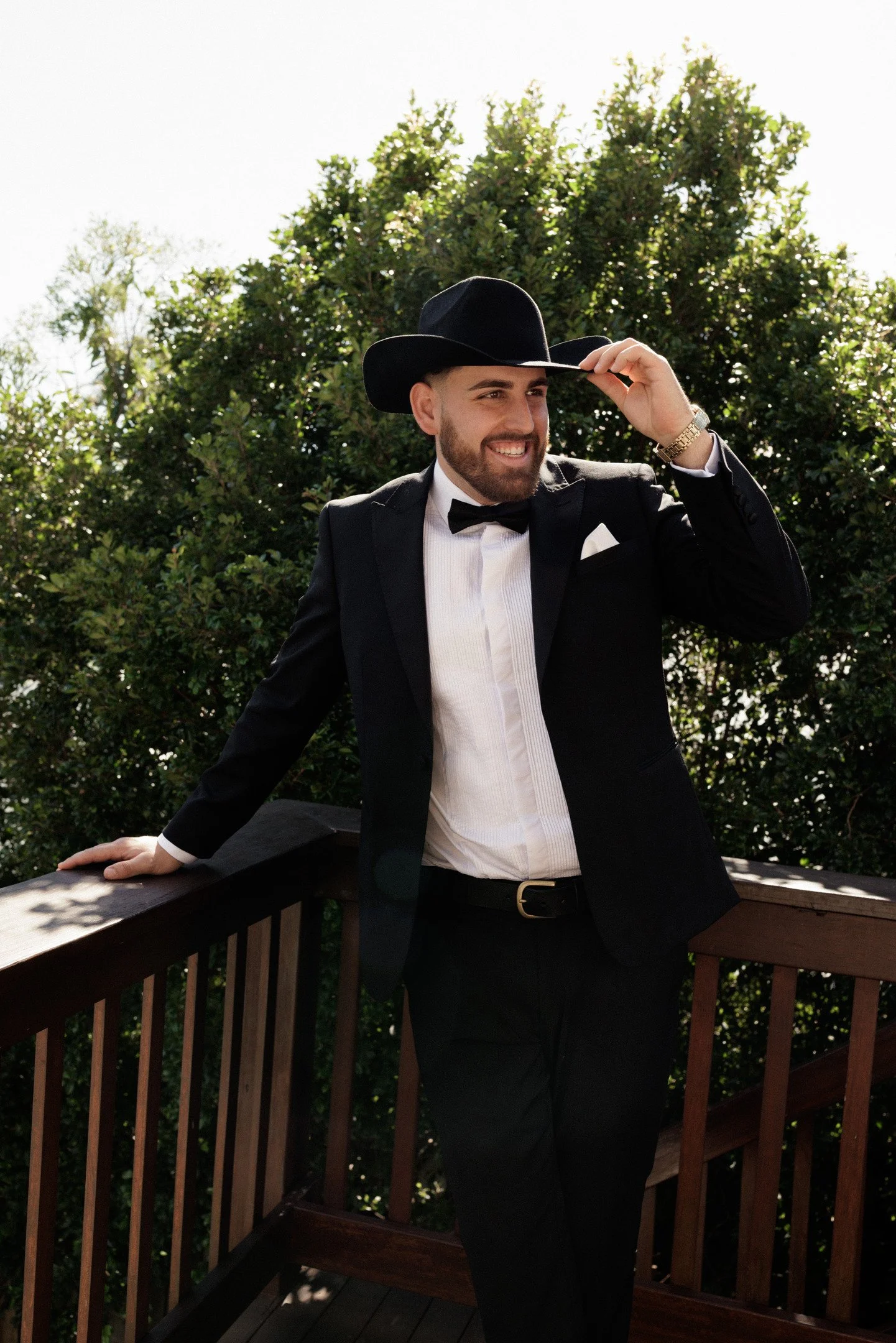 A man dressed in a tuxedo and wearing a black cowboy hat, standing on a wooden deck with a railing, smiling and adjusting his hat, with greenery and trees in the background.