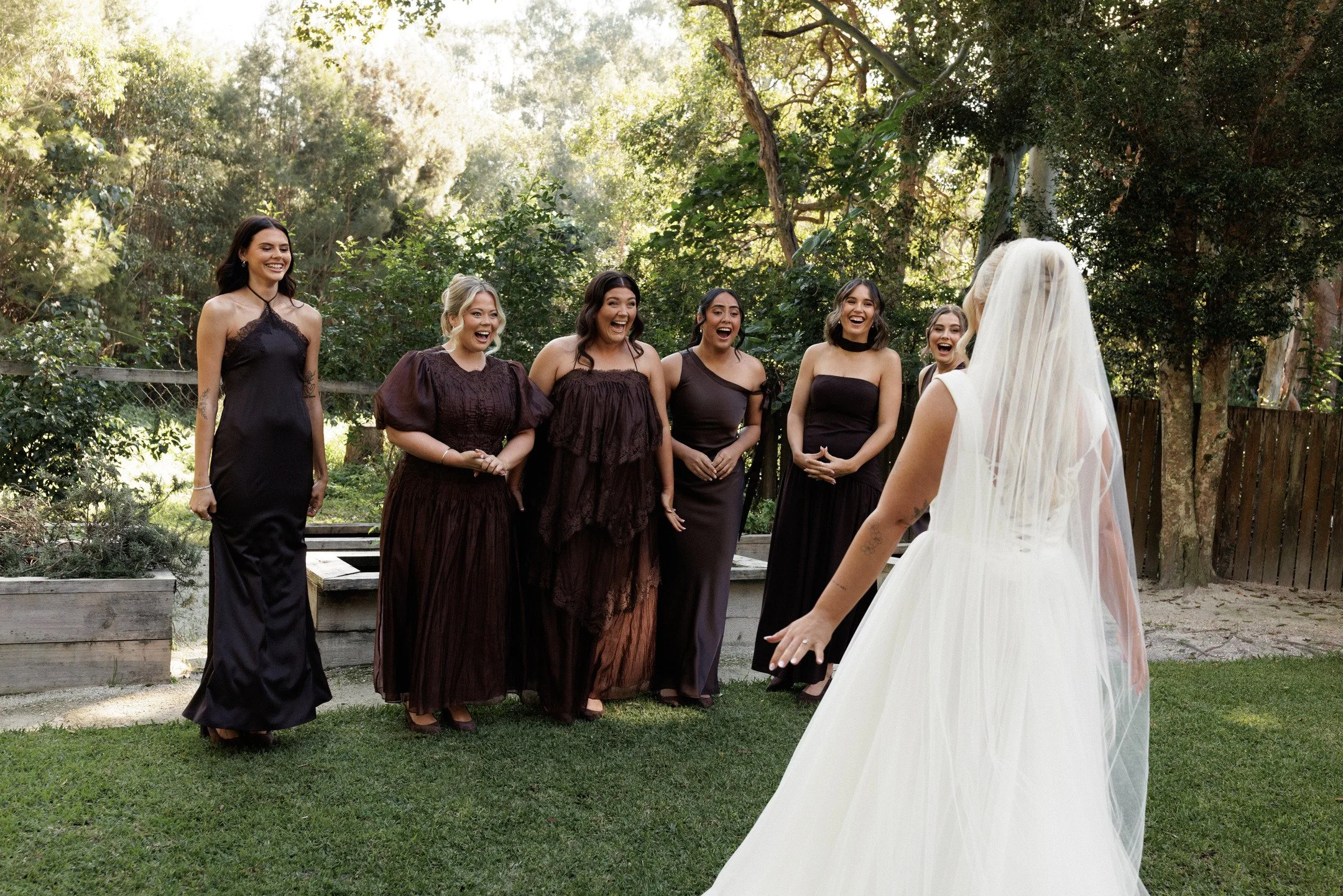 A bride in a white wedding dress with a veil is facing a group of six women in black dresses, who are smiling and laughing outdoors in a lush, wooded area.
