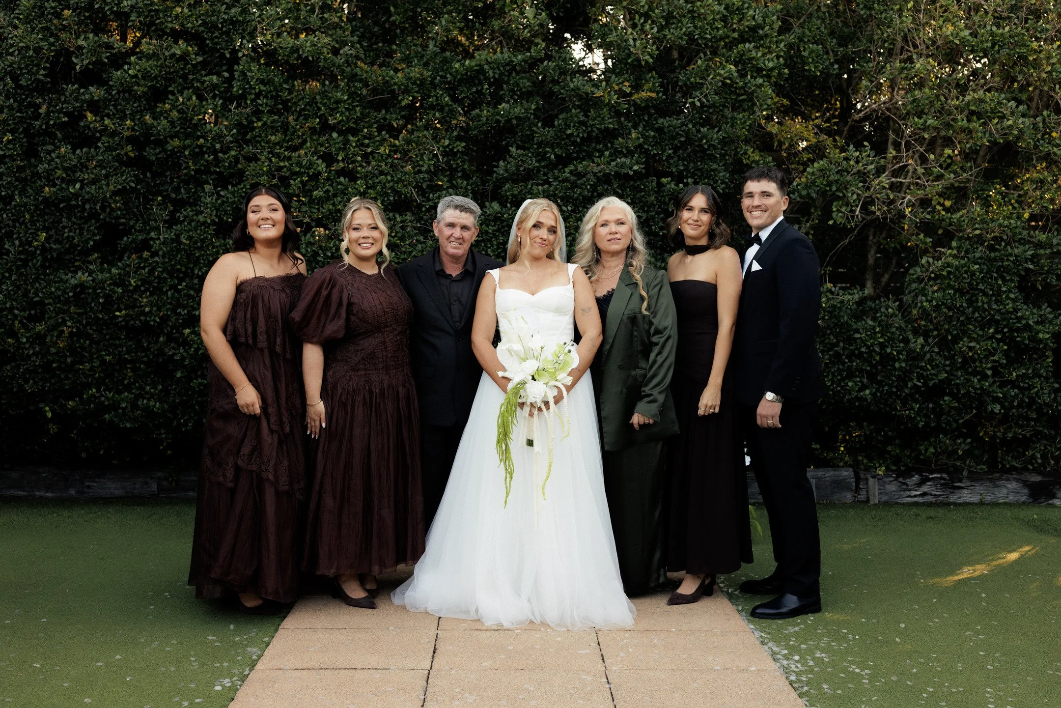 A group of seven people, dressed formally, standing outdoors in front of a lush green hedge. The group includes five women and two men, with one woman in a white wedding dress holding a bouquet. The others are wearing elegant dresses and suits.