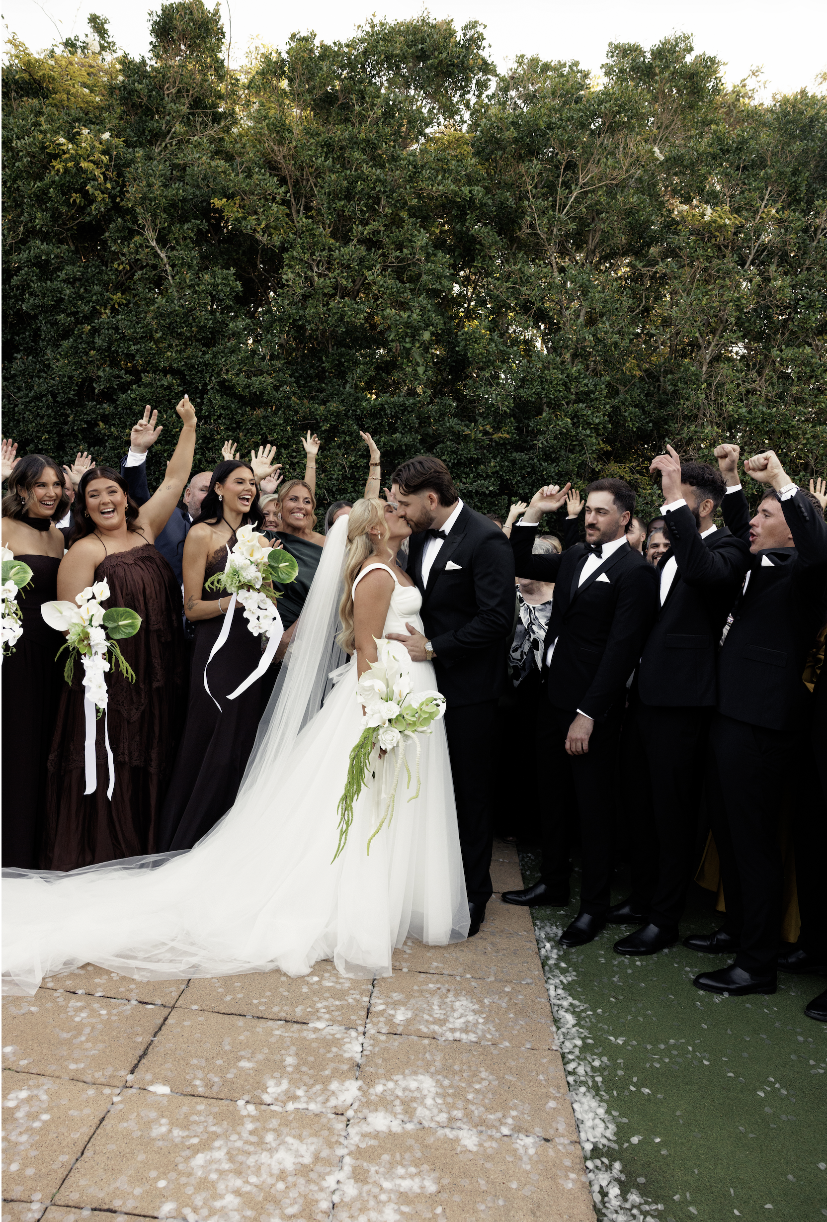 A bride and groom kiss at their outdoor wedding ceremony, surrounded by guests dressed in formal attire, with white flower petals on the ground and green trees in the background.