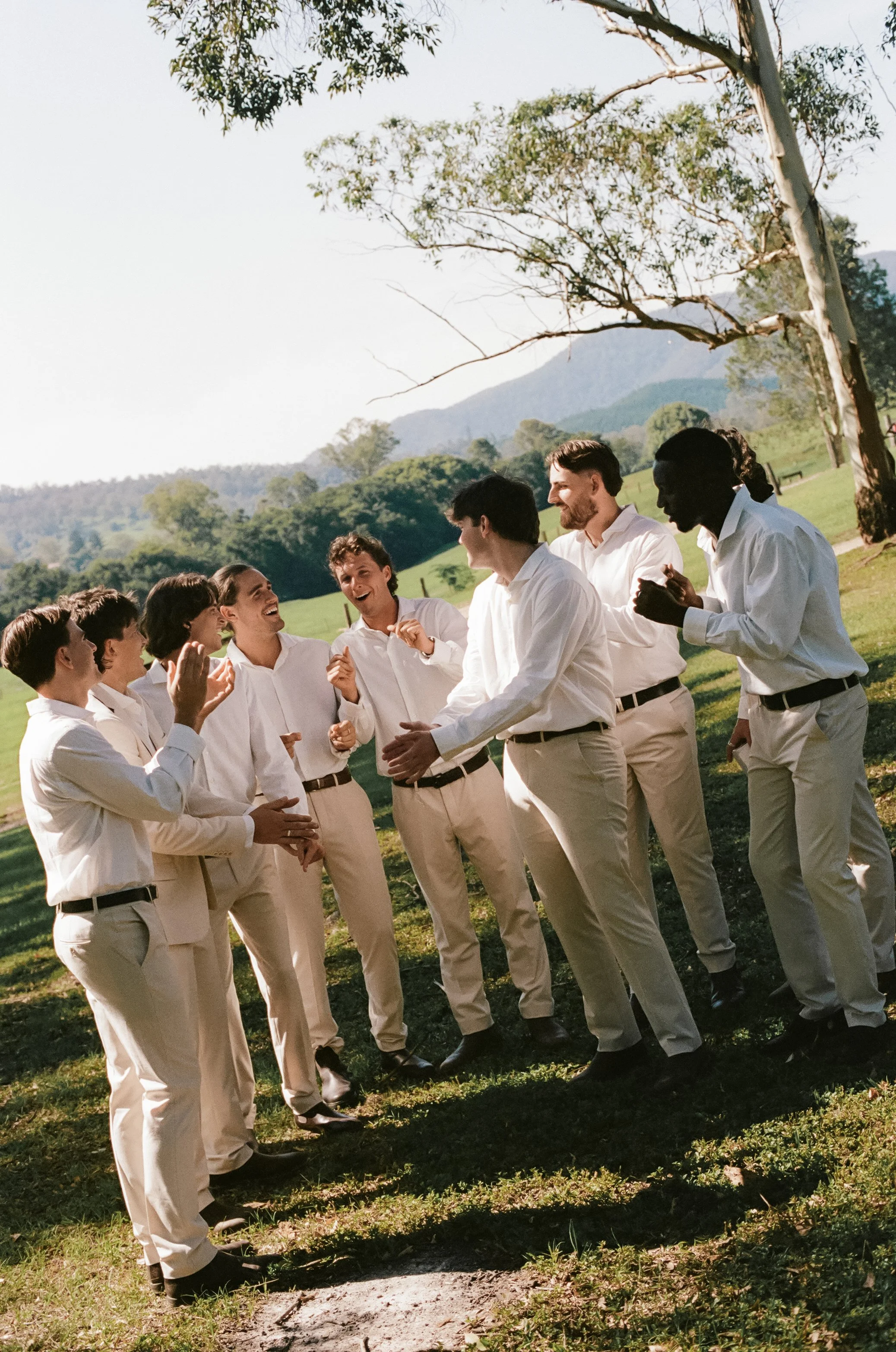 Group of people dressed in white shirts and light-colored pants standing outdoors on a sunny day, engaged in conversation and smiling.