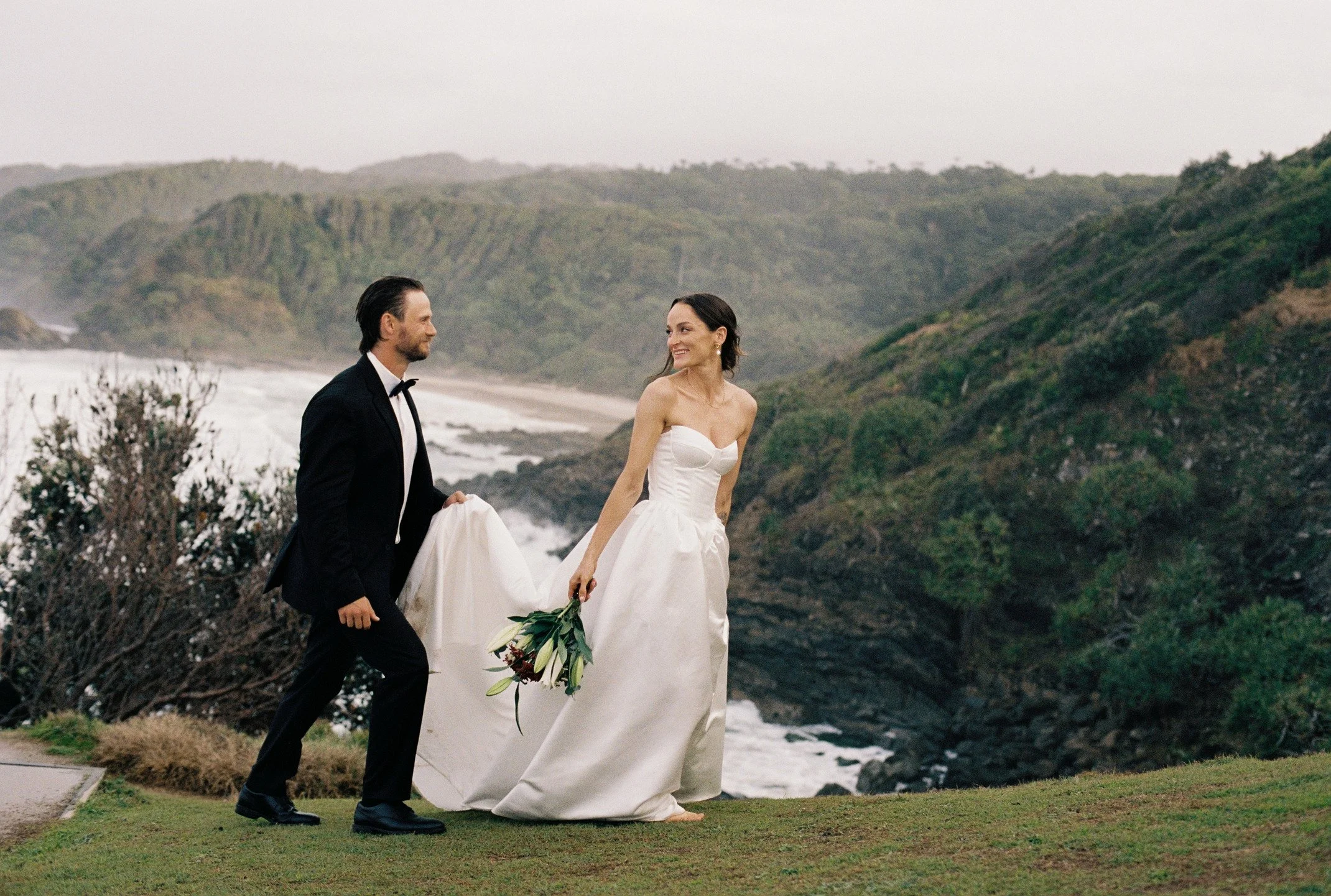 A bride and groom walking along a grassy area near the coast with cliffs and ocean in the background, during a cloudy day.