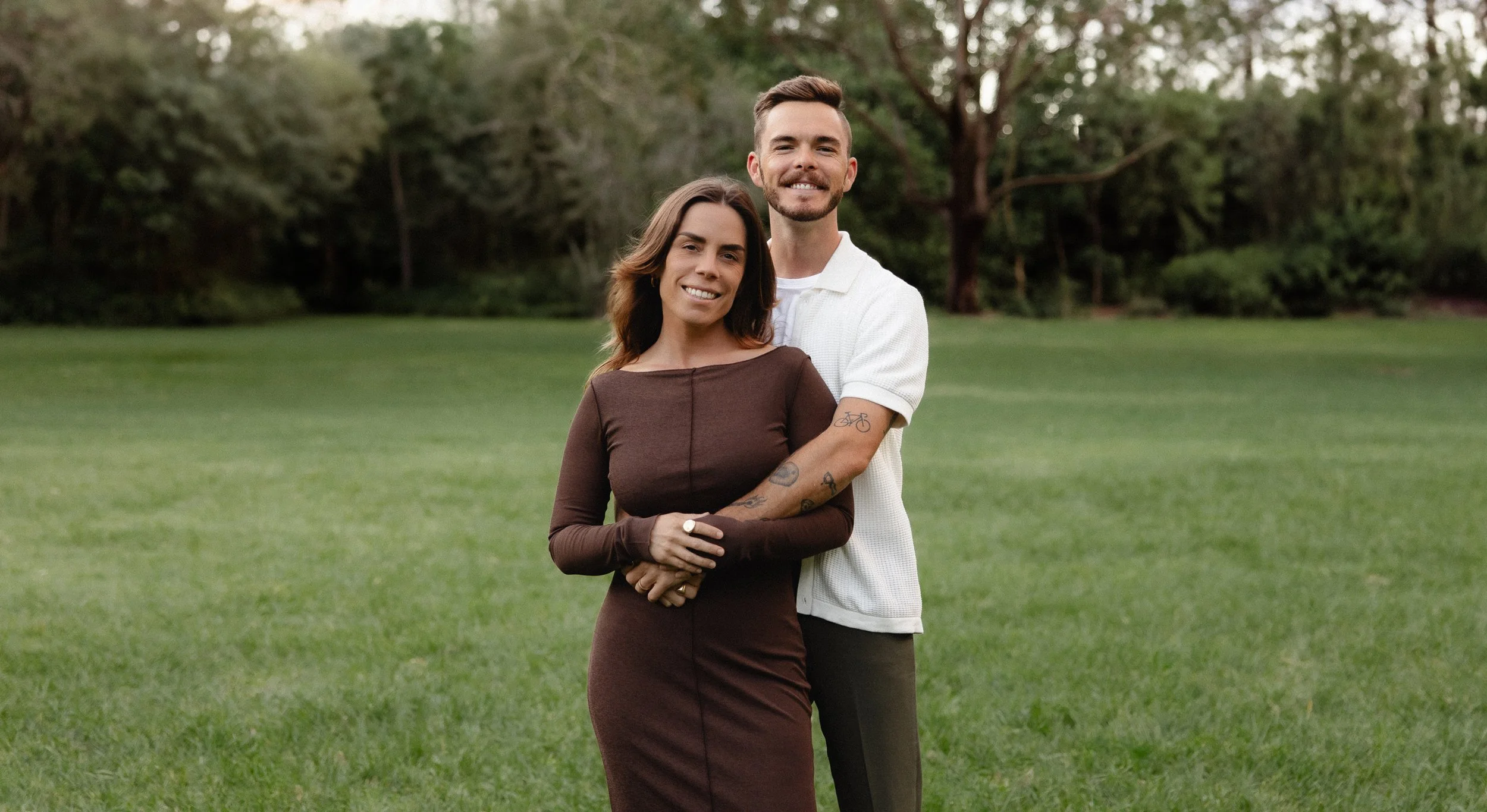 A smiling couple standing in a grassy park with trees in the background, with the man hugging the woman from behind.