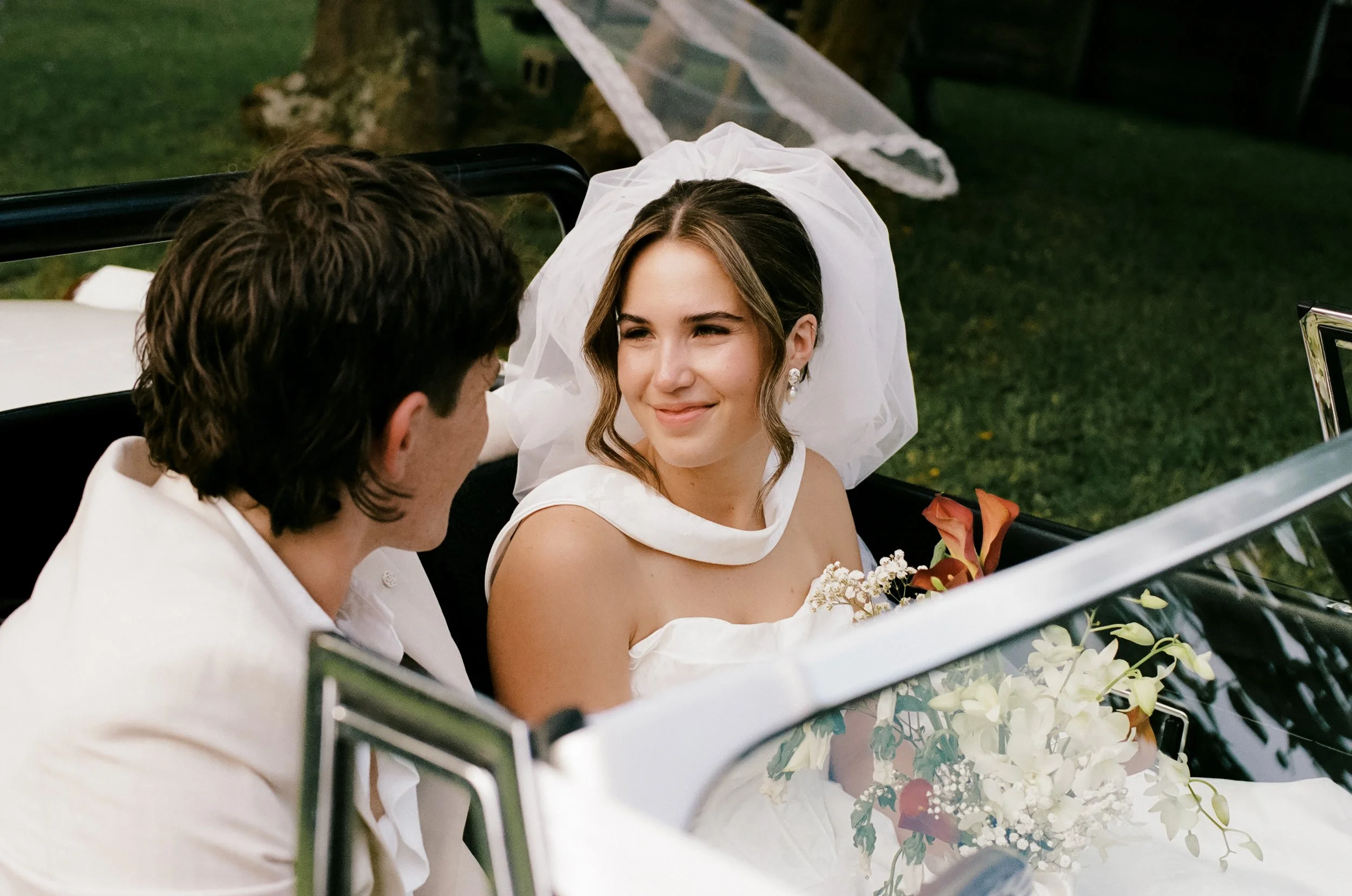 Bride and groom sitting in a vintage car, smiling at each other during wedding.