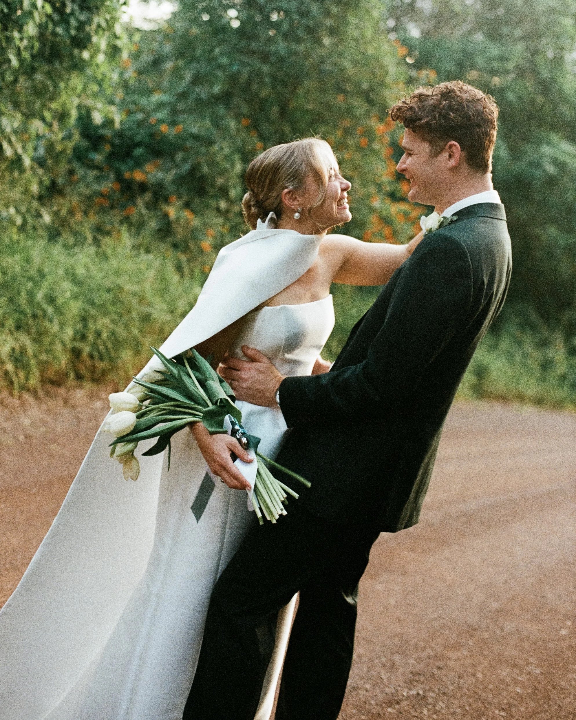 A bride and groom smiling and embracing outdoors during sunset, with greenery in the background. The bride is holding a bouquet of white tulips.