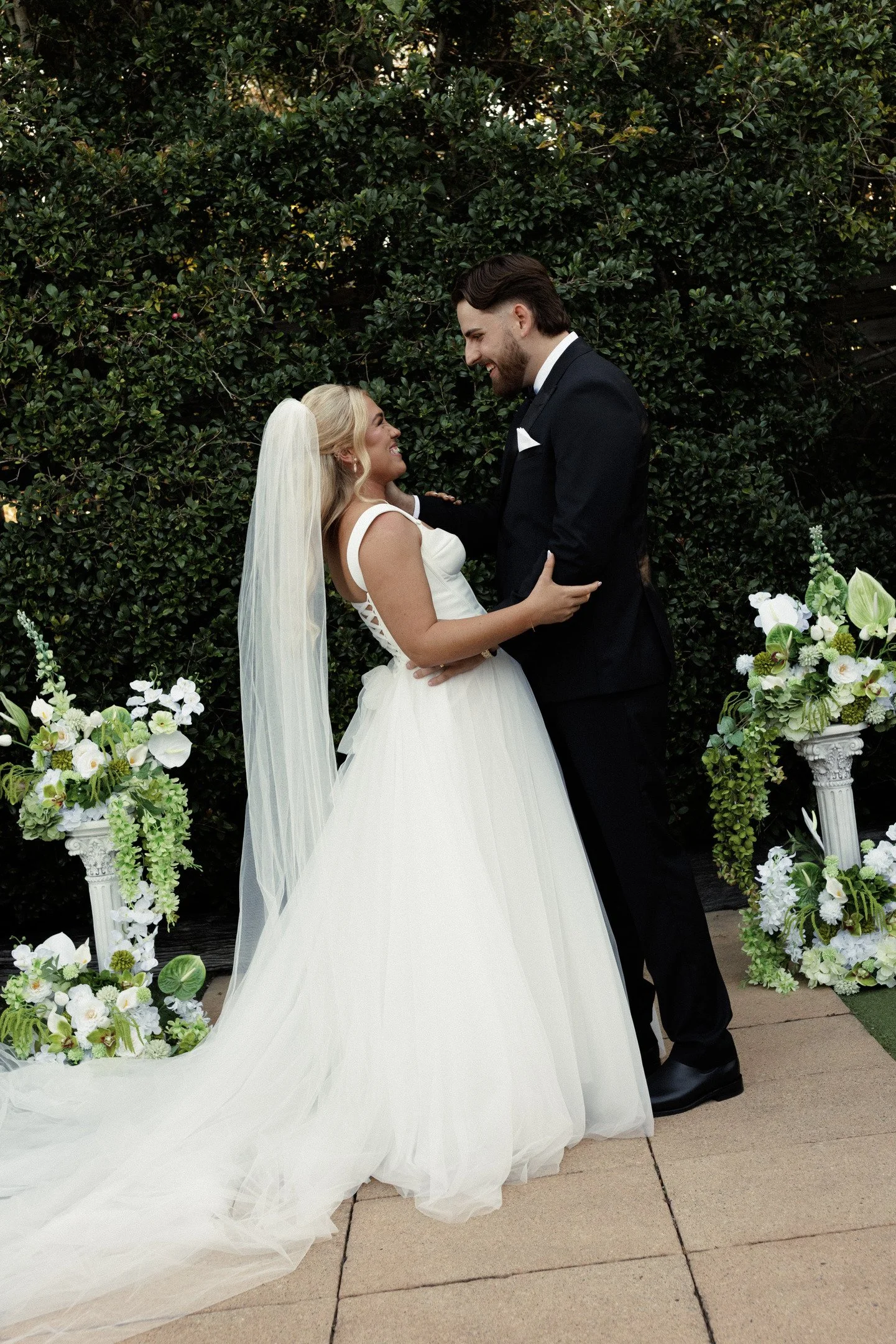 A bride and groom standing outdoors in front of a green hedge, smiling at each other, with floral arrangements on either side.