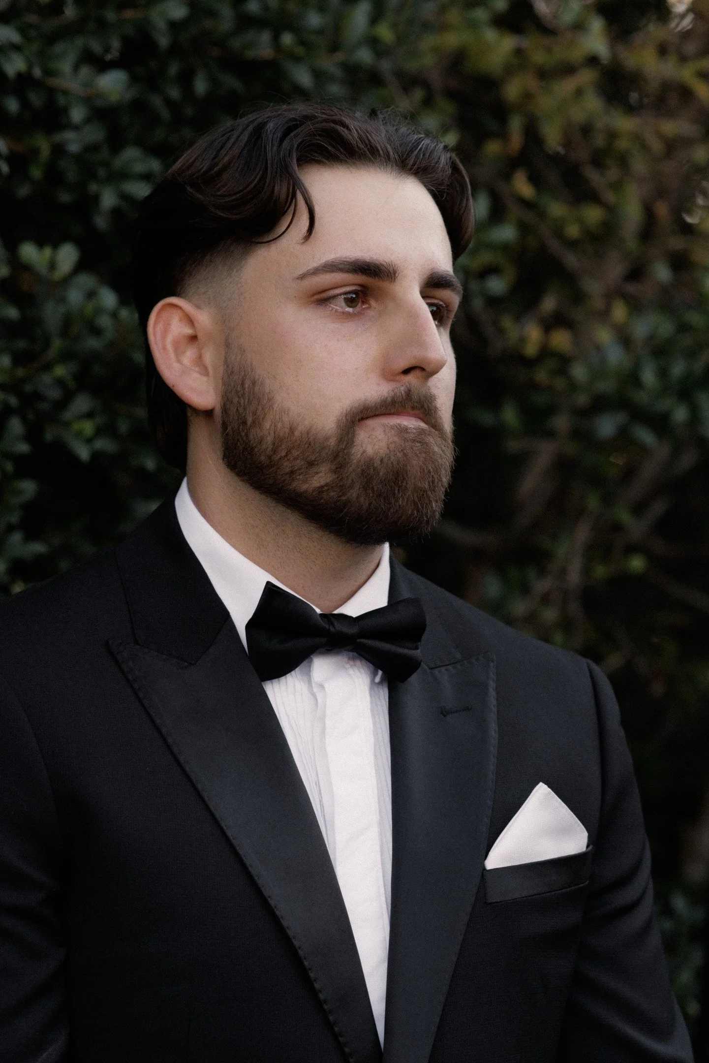 A man with brown hair and a beard dressed in a black tuxedo, white dress shirt, black bow tie, and white pocket square, standing outdoors in front of greenery.