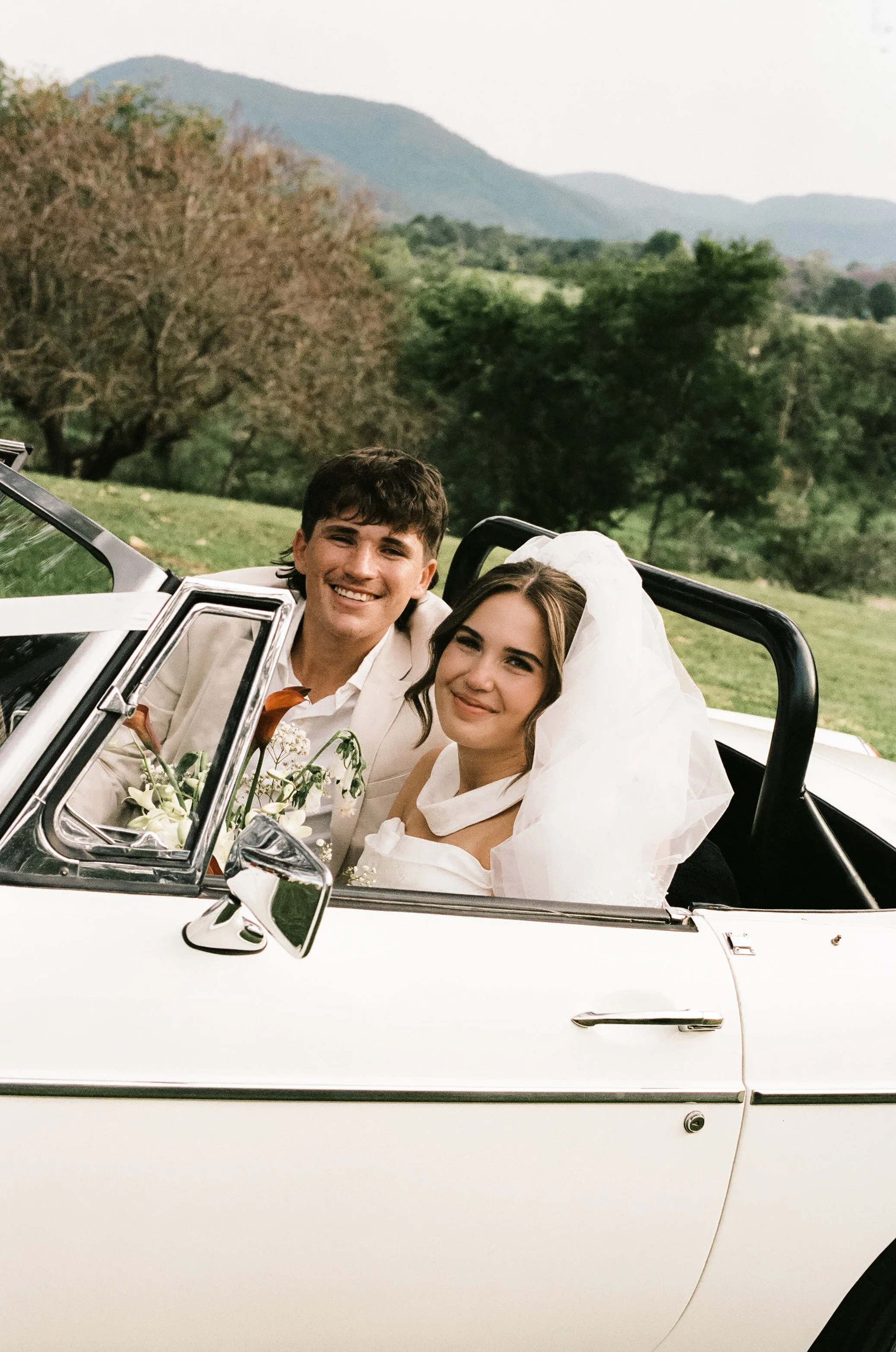 A newlywed couple in wedding attire smiling and sitting in a white convertible car with a scenic outdoor background of trees, mountains, and greenery.