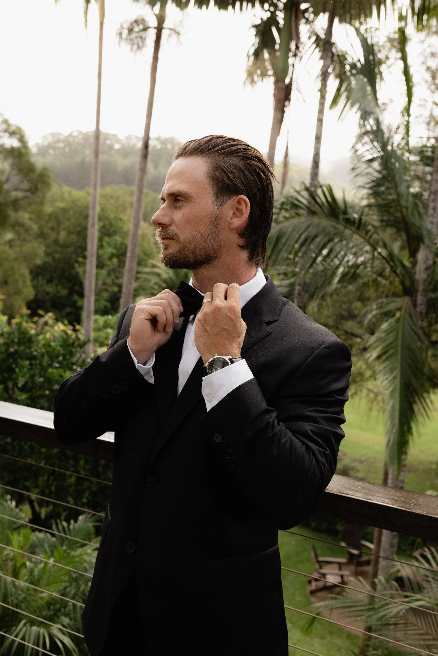 A man in a black tuxedo adjusting his bow tie outdoors with tropical trees and greenery in the background.