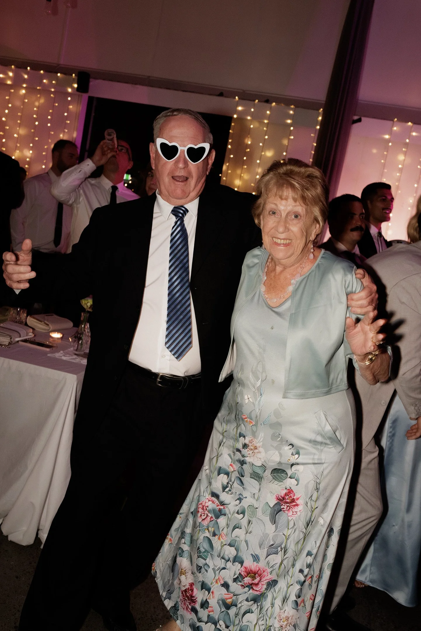 A man and an elderly woman dressed in formal attire celebrating at a party, with string lights and other guests in the background.