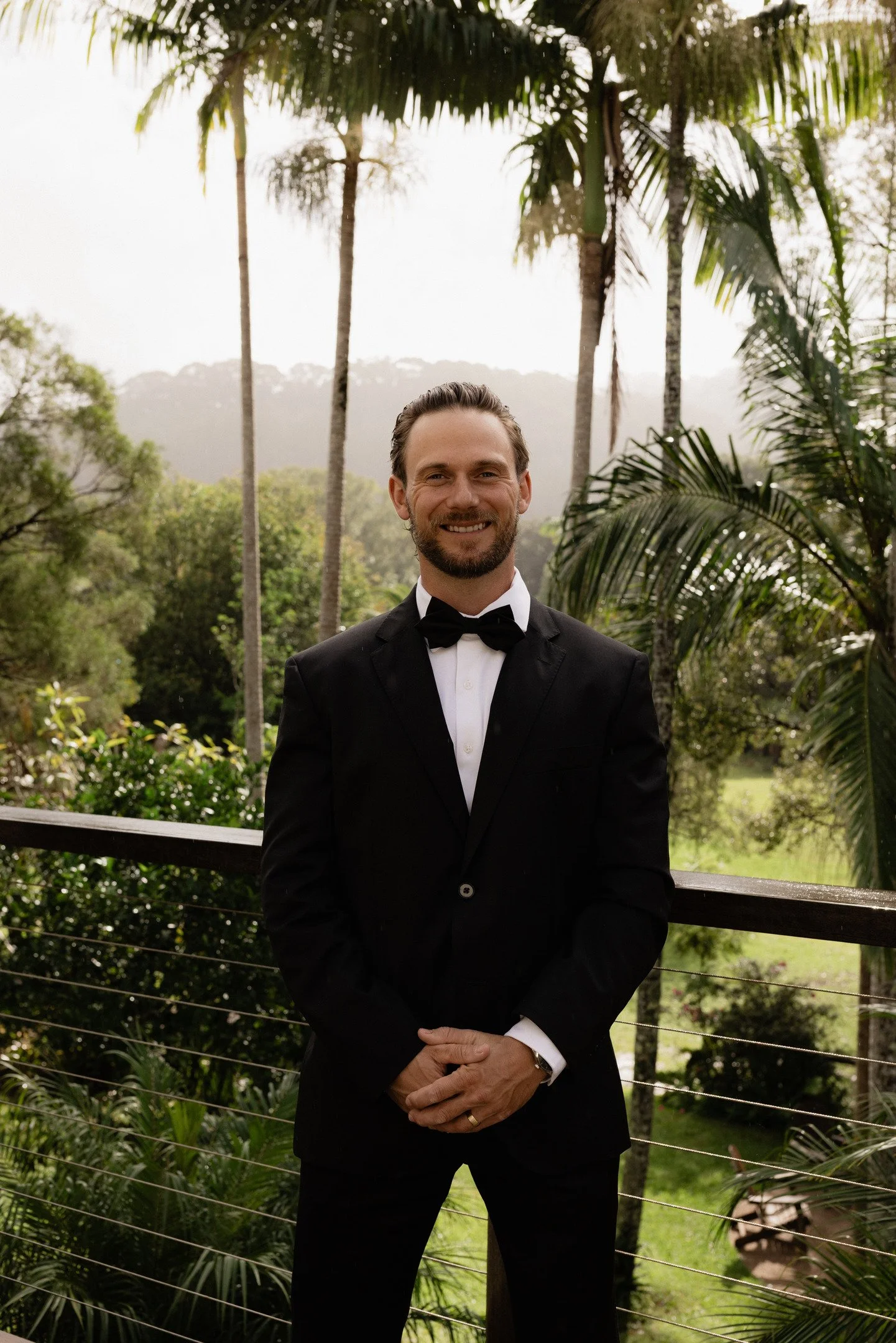 A man in a black tuxedo with a white shirt and black bow tie standing outdoors in front of green trees and palm plants, smiling at the camera.