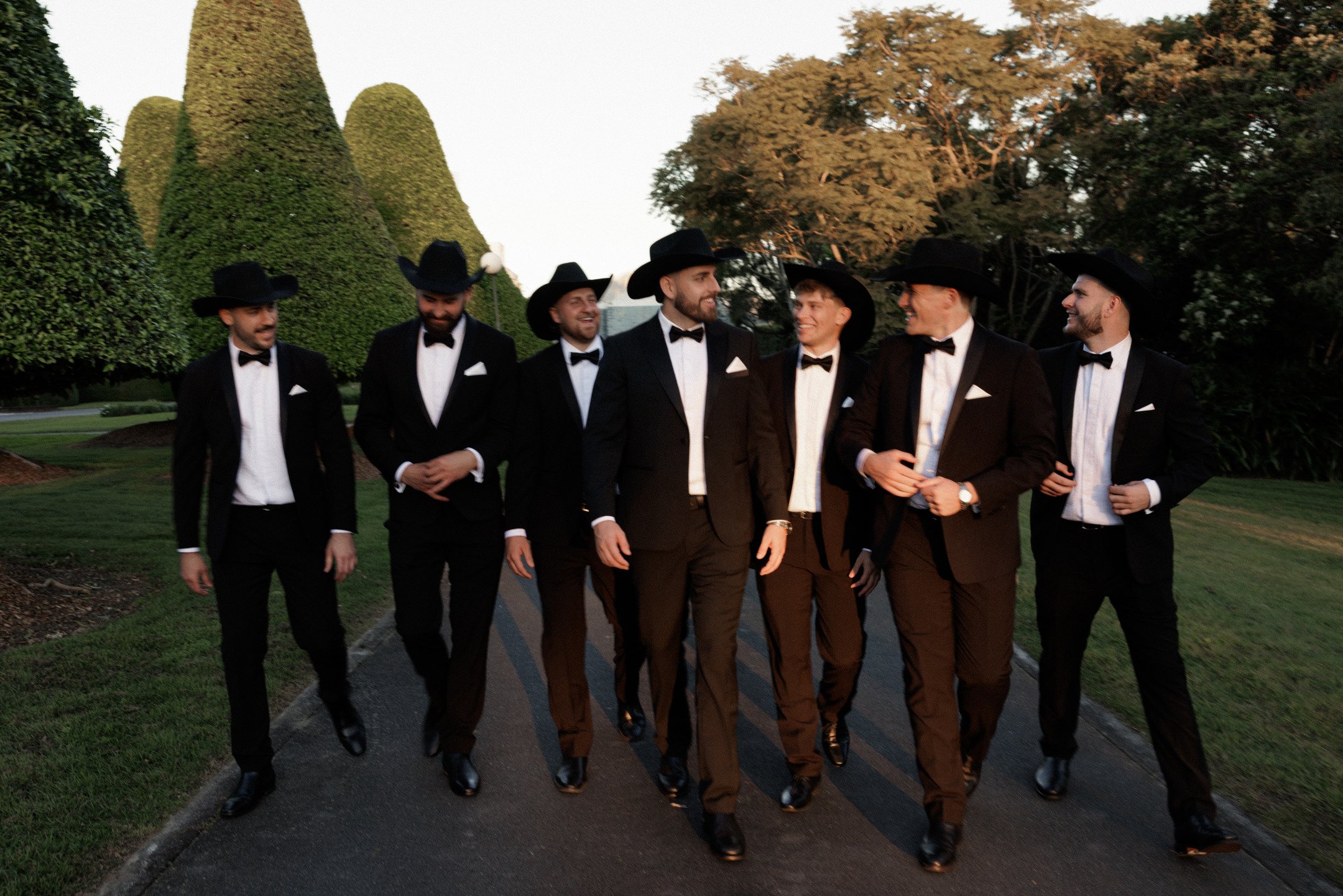 A group of seven men in tuxedos and cowboy hats walking and smiling outdoors in a park with large trimmed bushes and trees.