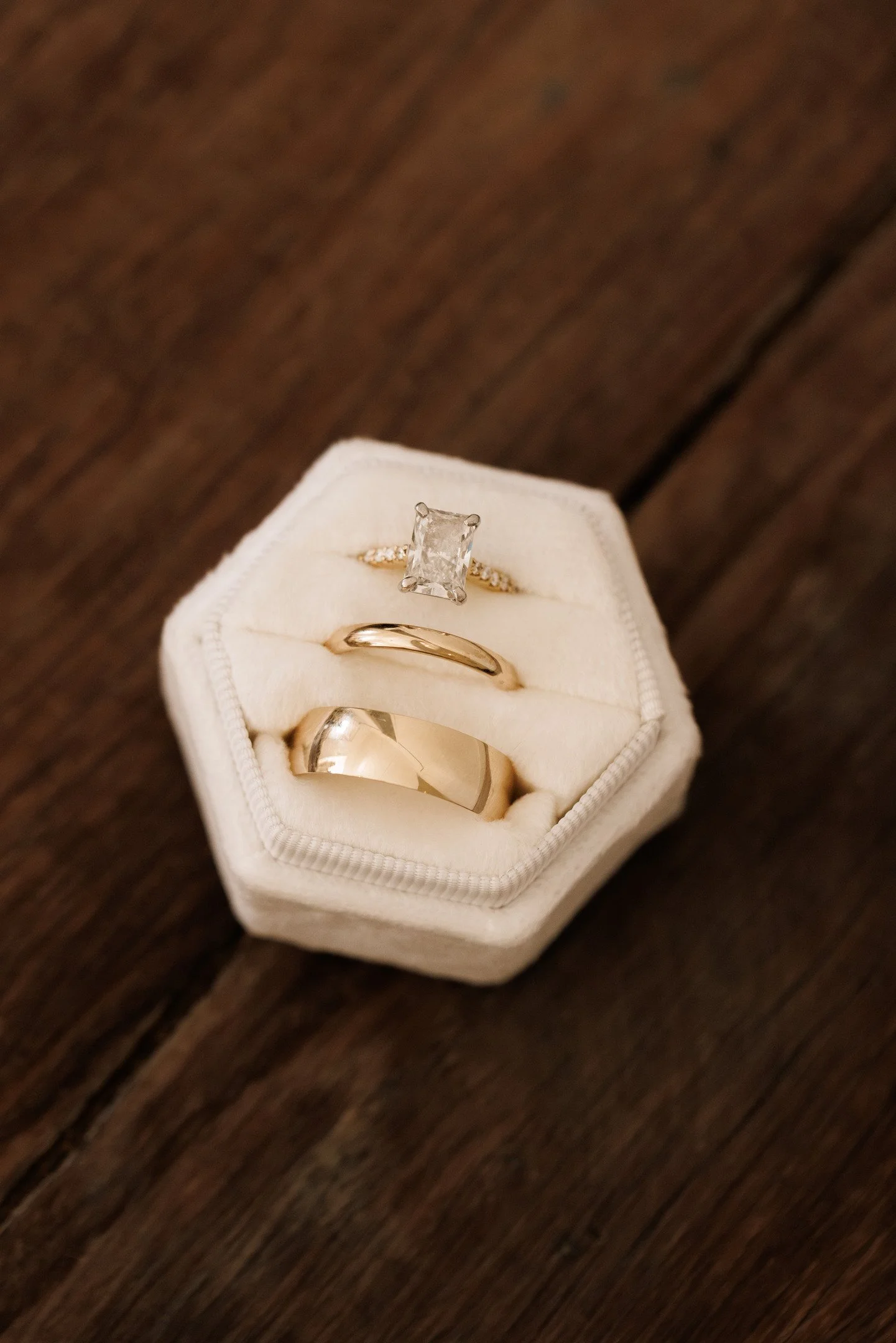 A white jewelry box holding three rings: a large rectangular diamond ring, a gold band, and a silver ring, on a wooden surface.