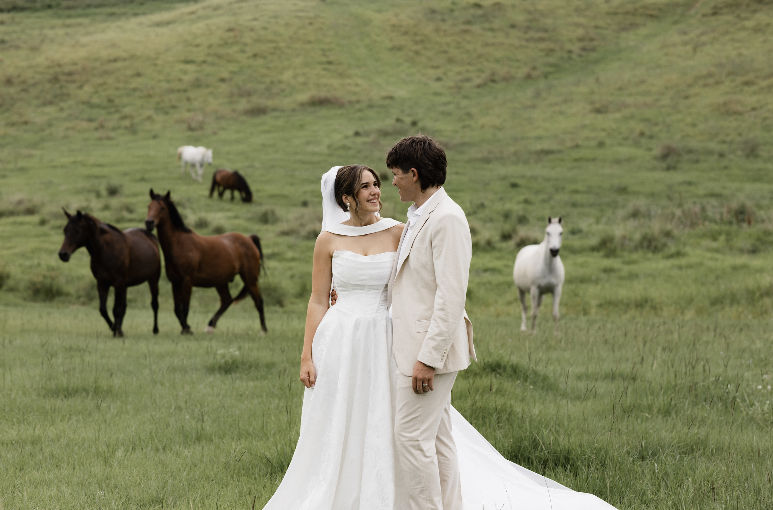 A bride and groom stand together on green grass, smiling at each other, with horses grazing in the background on a hillside.
