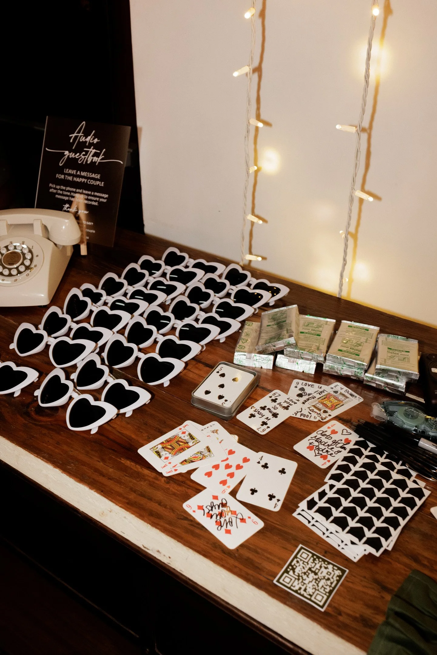 Table with scattered playing cards, black heart-shaped sunglasses, small stacks of wrapped gum, and a sign that reads 'Audio Guestbook' with instructions to leave a message for the happy couple. There is also a vintage rotary phone and a QR code on t