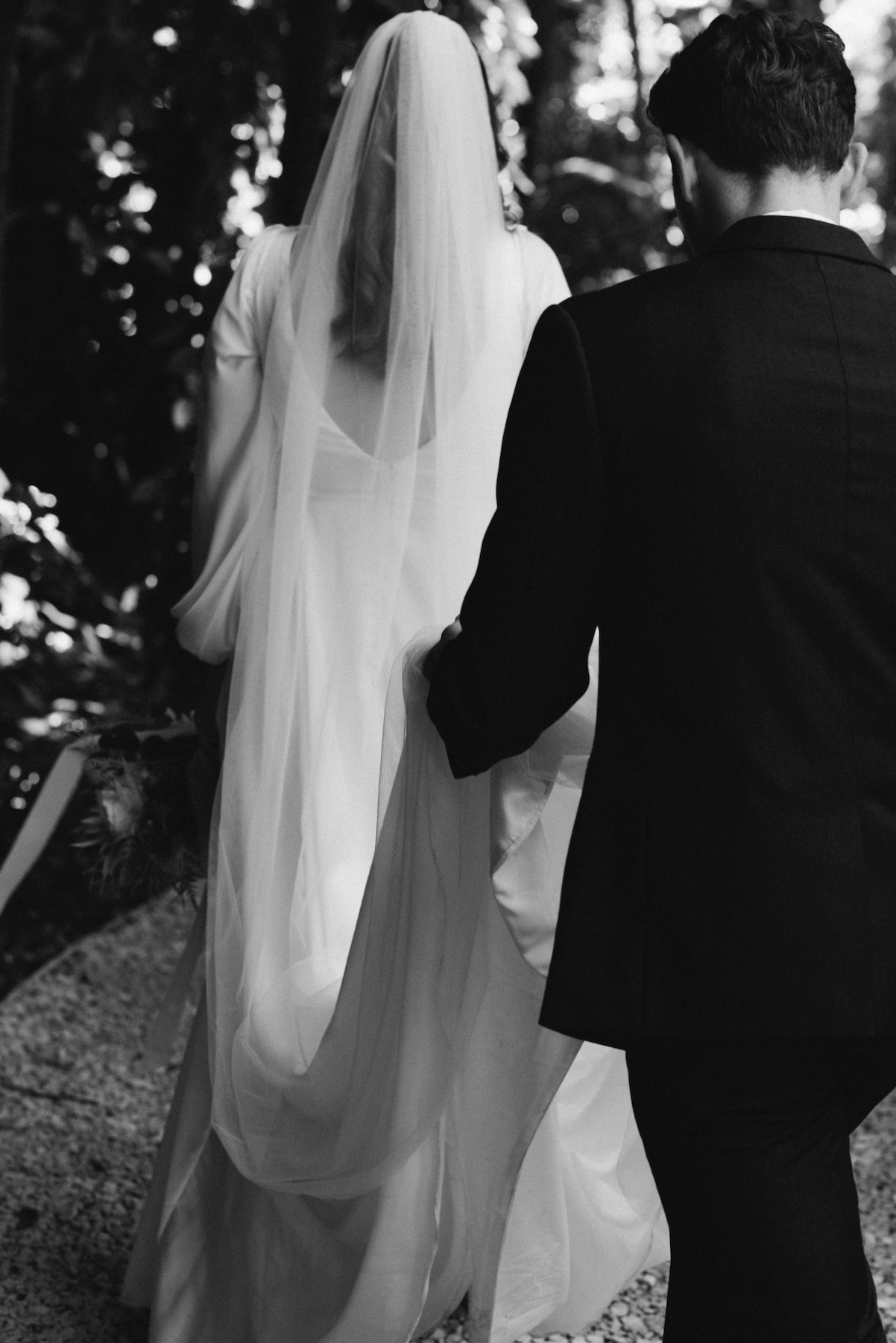 A black and white photograph of a bride and groom walking outdoors during their wedding. The bride is wearing a long flowing wedding gown and veil, and the groom is dressed in a dark suit. They are holding hands, and trees are visible in the backgrou