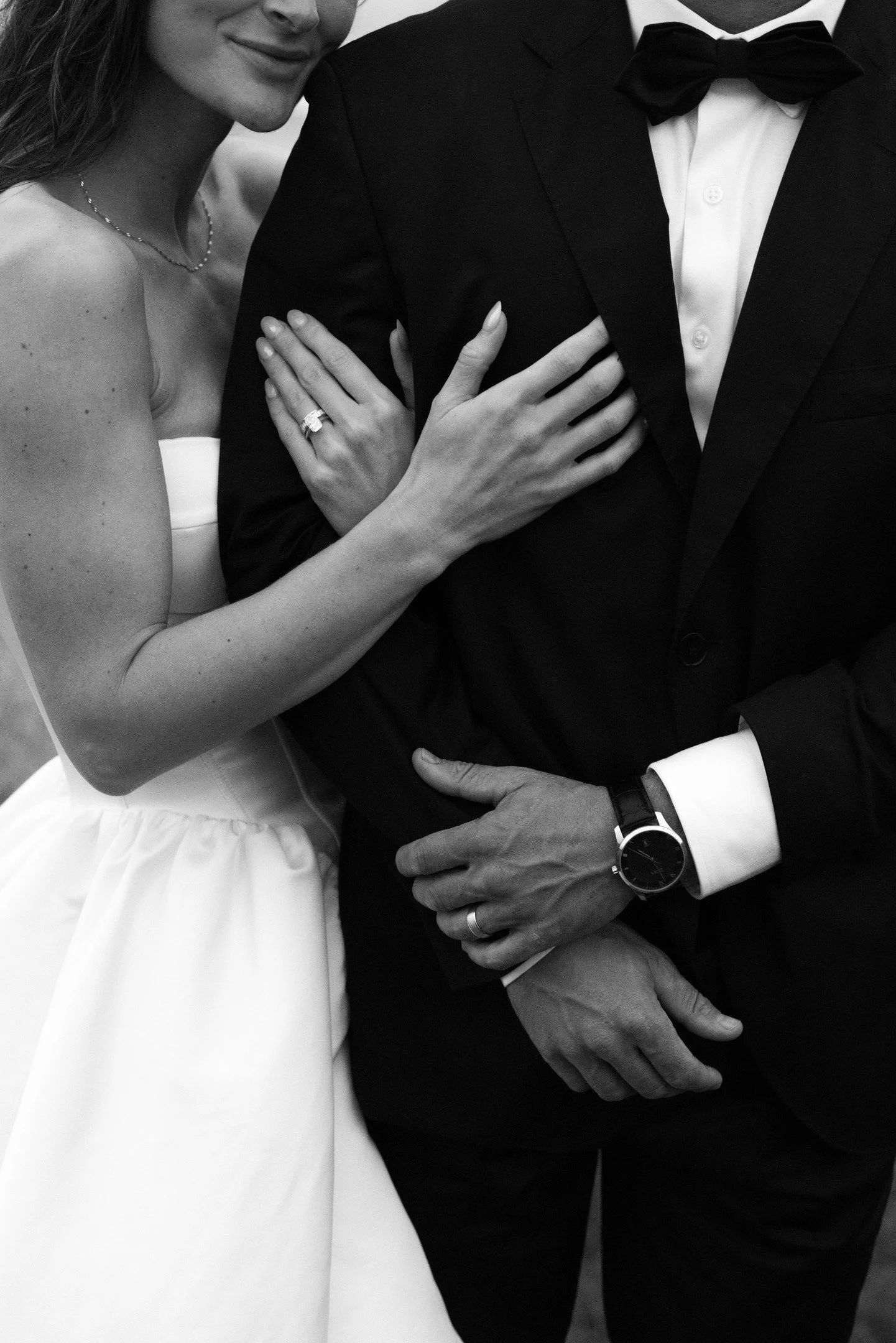 Close-up black and white photo of a bride and groom. The bride's hand, showing a wedding ring, rests on the groom's arm. The bride is wearing a strapless wedding dress and a delicate necklace. The groom is dressed in a tuxedo with a bow tie, and his 